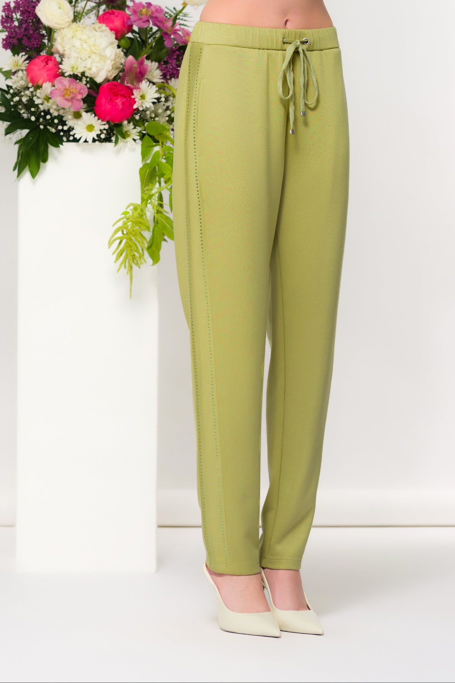Women’s slim ankle-length pants with a streamlined fit and drawstring waist, styled with a cropped long-sleeve top and heels in a studio setting with floral accents.