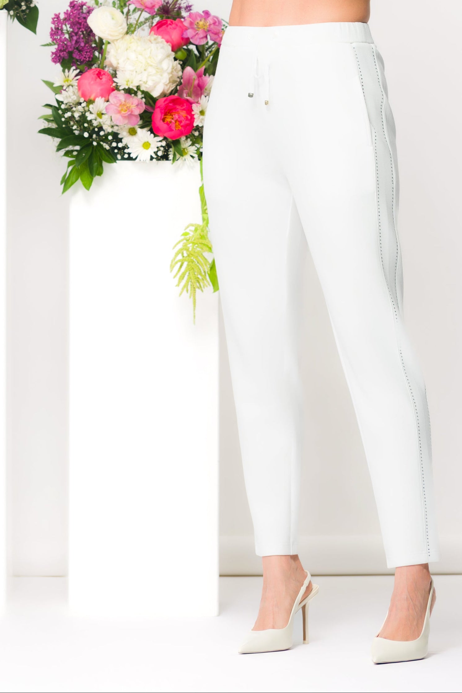 Women’s slim ankle-length pants with a streamlined fit and drawstring waist, styled with a cropped long-sleeve top and heels in a studio setting with floral accents.