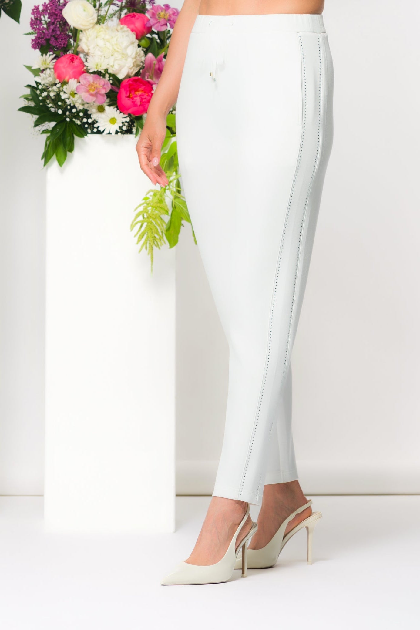 Women’s slim ankle-length pants with a streamlined fit and drawstring waist, styled with a cropped long-sleeve top and heels in a studio setting with floral accents.