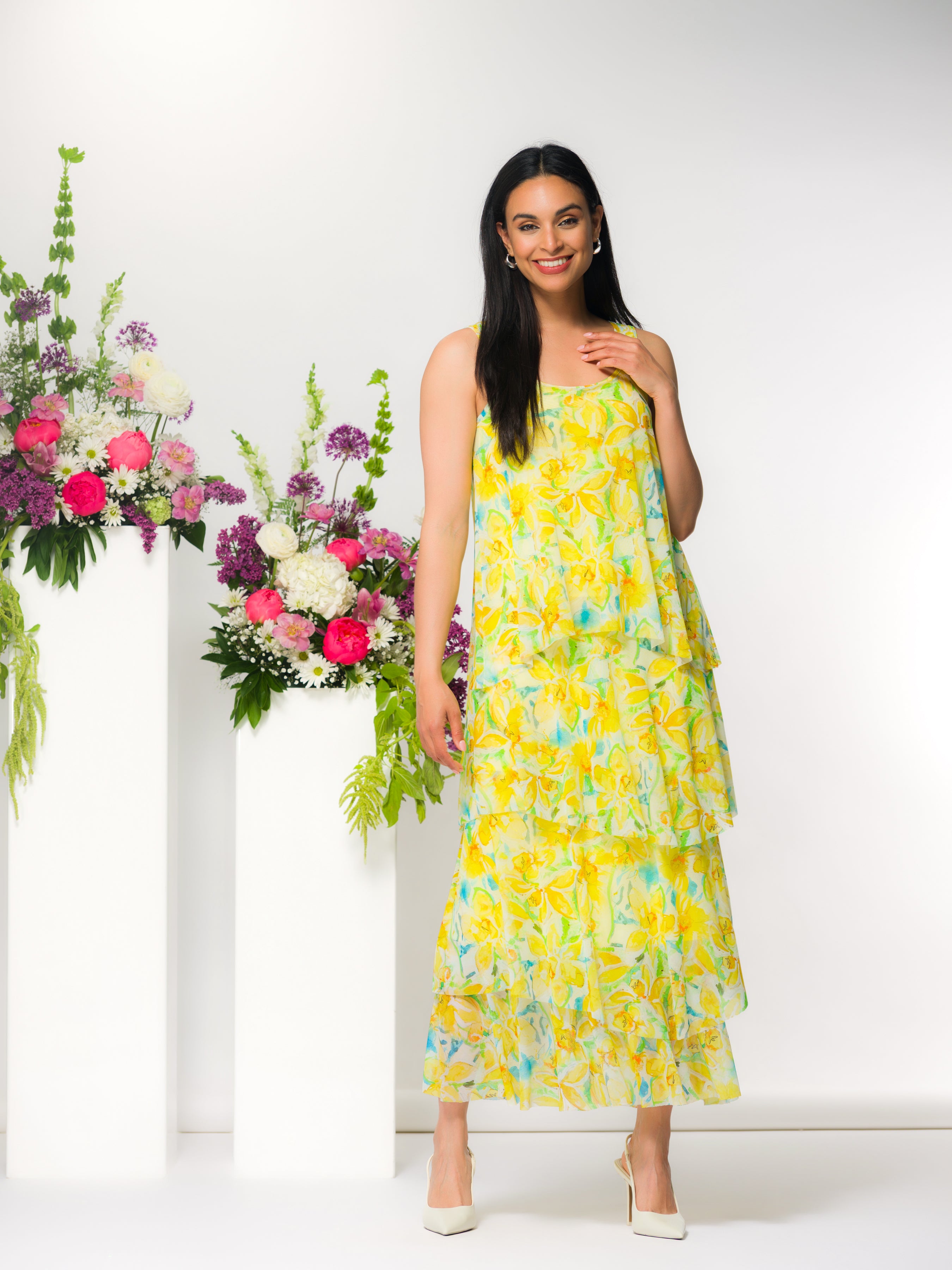 Model wearing a sleeveless tiered midi dress with a flowing silhouette, styled with heels, standing in a studio setting with floral arrangements on pedestal stands.