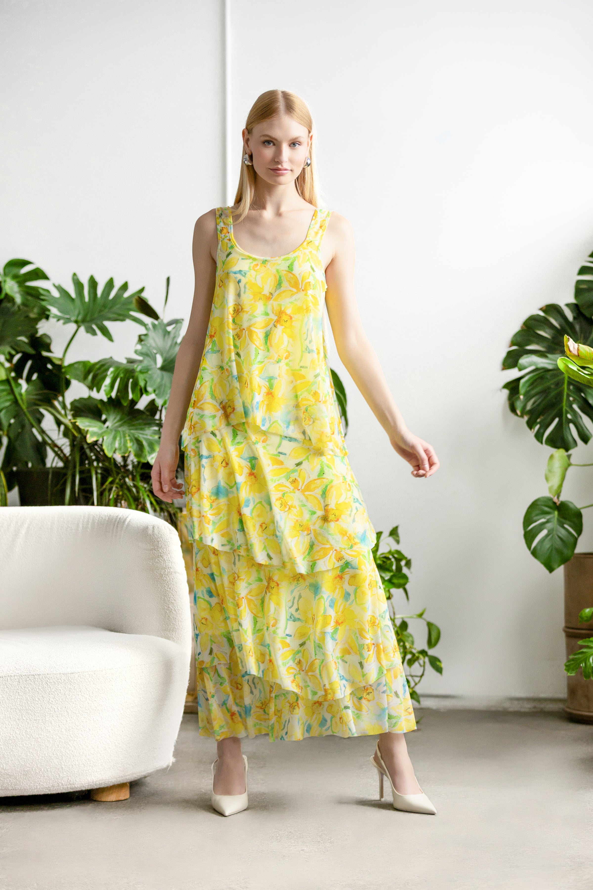 Model wearing a sleeveless tiered midi dress with a relaxed drape and scoop neckline, styled with heels, posed indoors among leafy plants and modern seating.