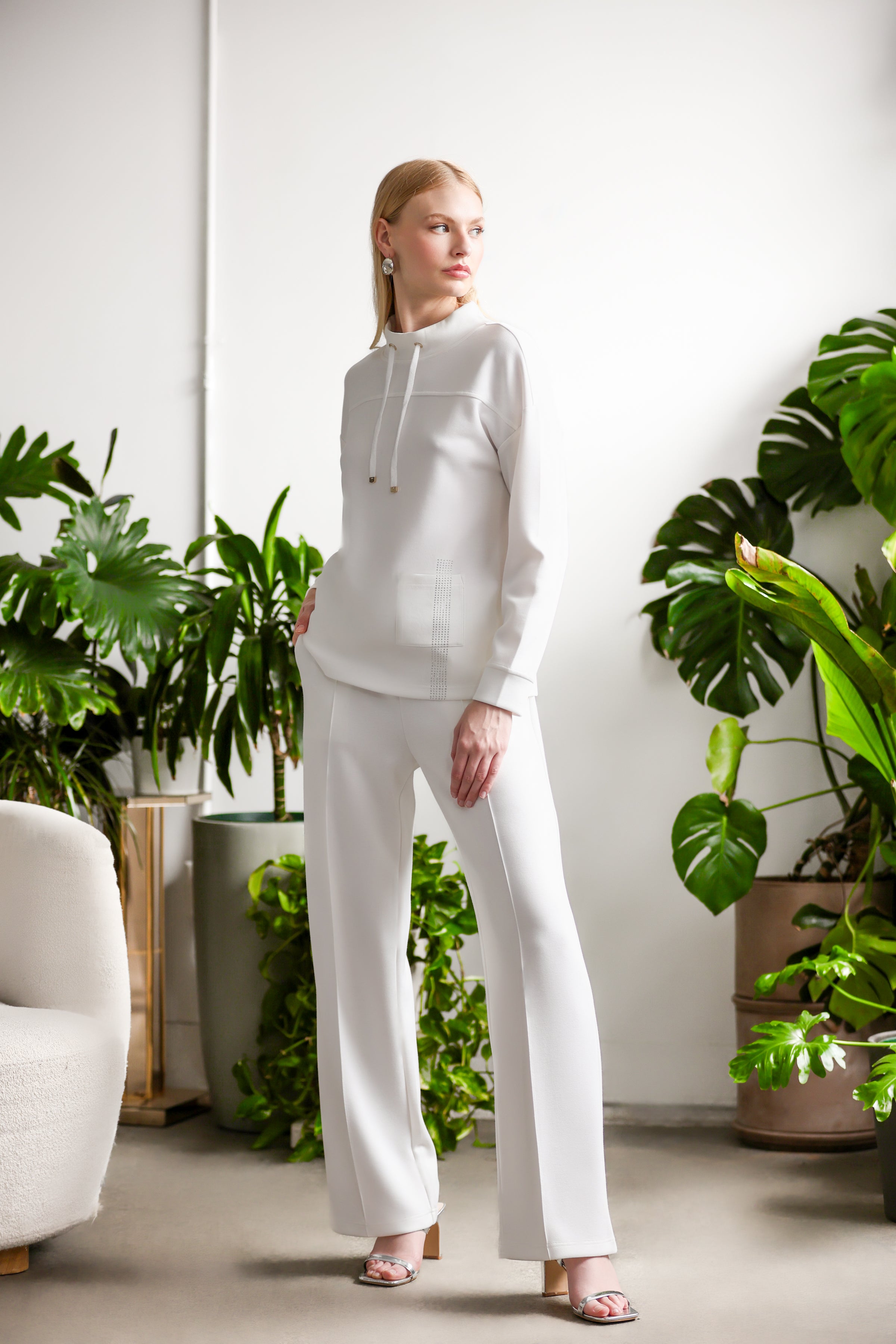 Model wearing a relaxed-fit pullover with long sleeves and straight-leg pants, styled with open-toe heels, standing in a bright interior space surrounded by indoor plants.