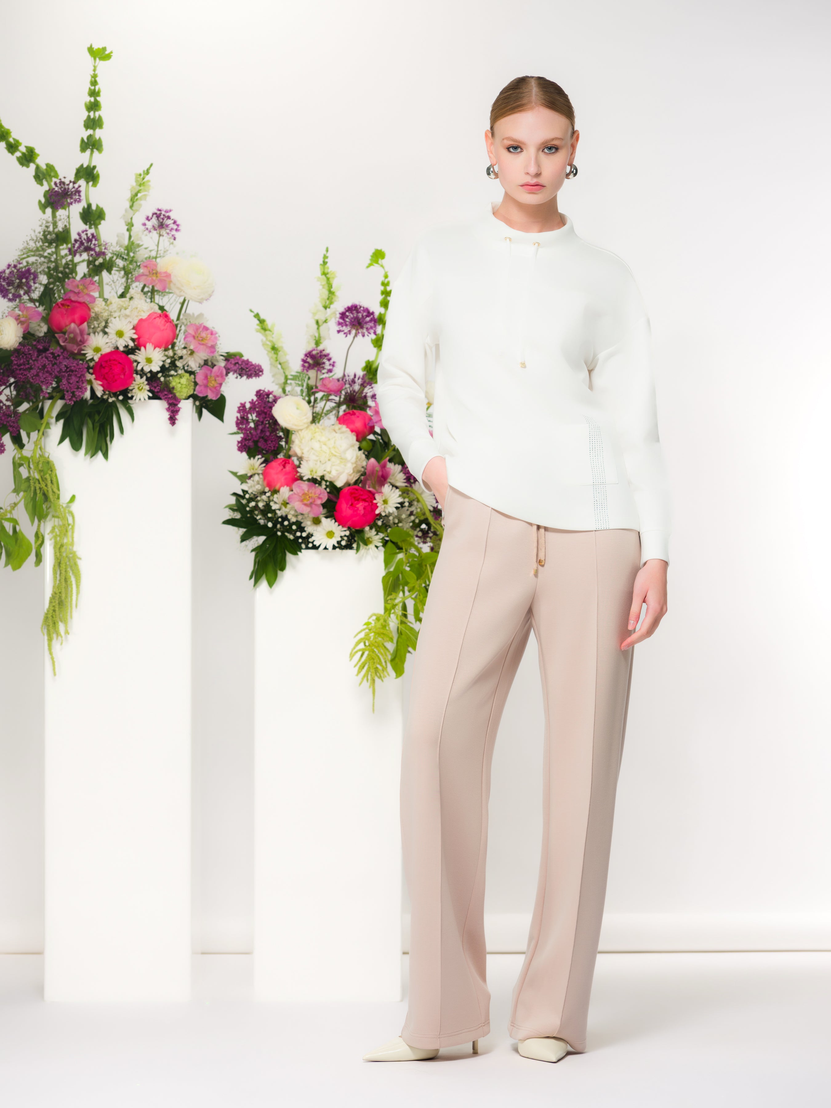 Model wearing a soft-drape pullover with a relaxed silhouette and straight-leg pants, styled with pointed-toe heels, photographed in a minimalist studio with floral arrangements.