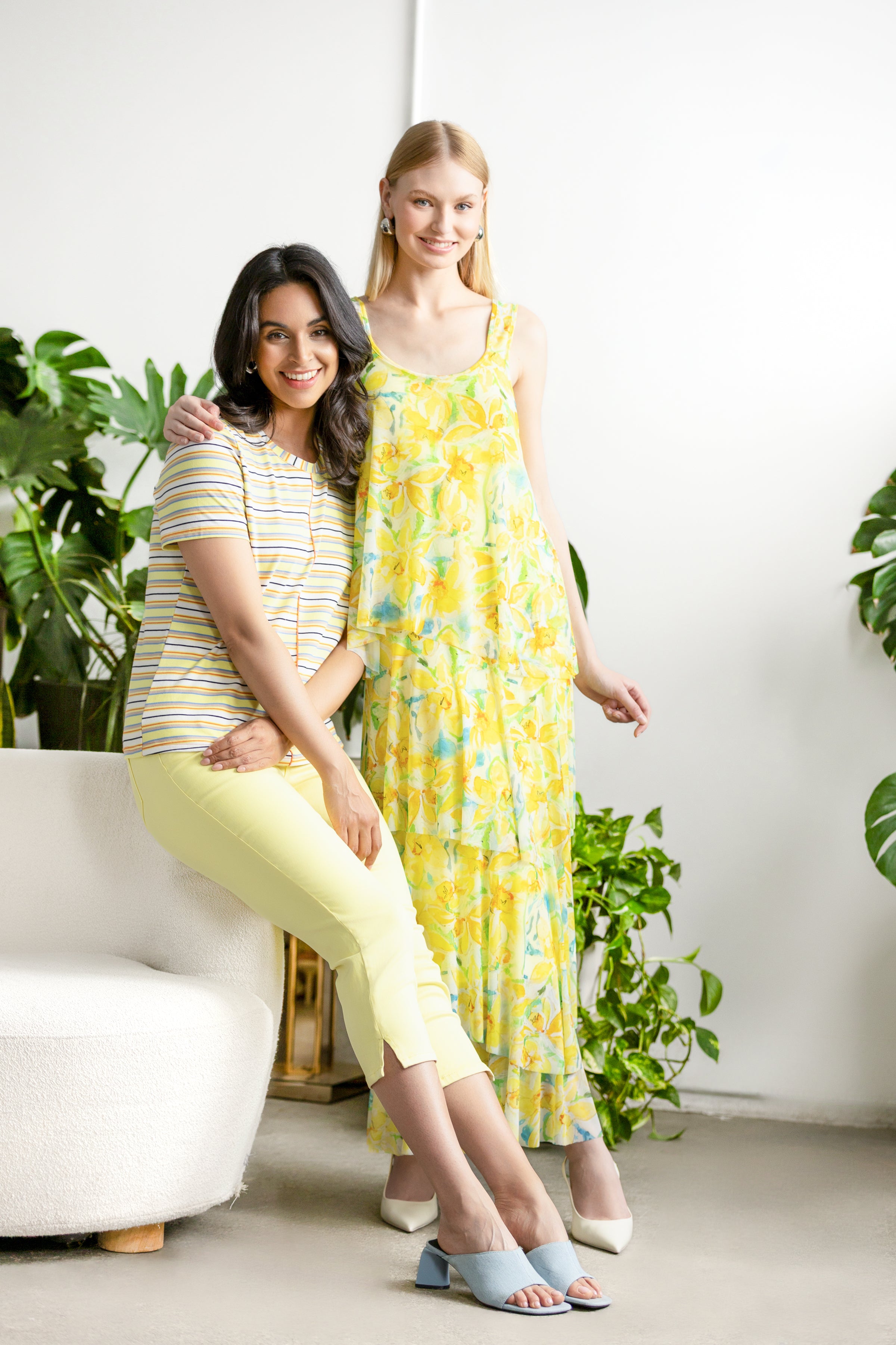 Two women wearing a relaxed-fit short-sleeve knit tee styled casually, one standing and one seated, photographed in a bright indoor setting with plants.
