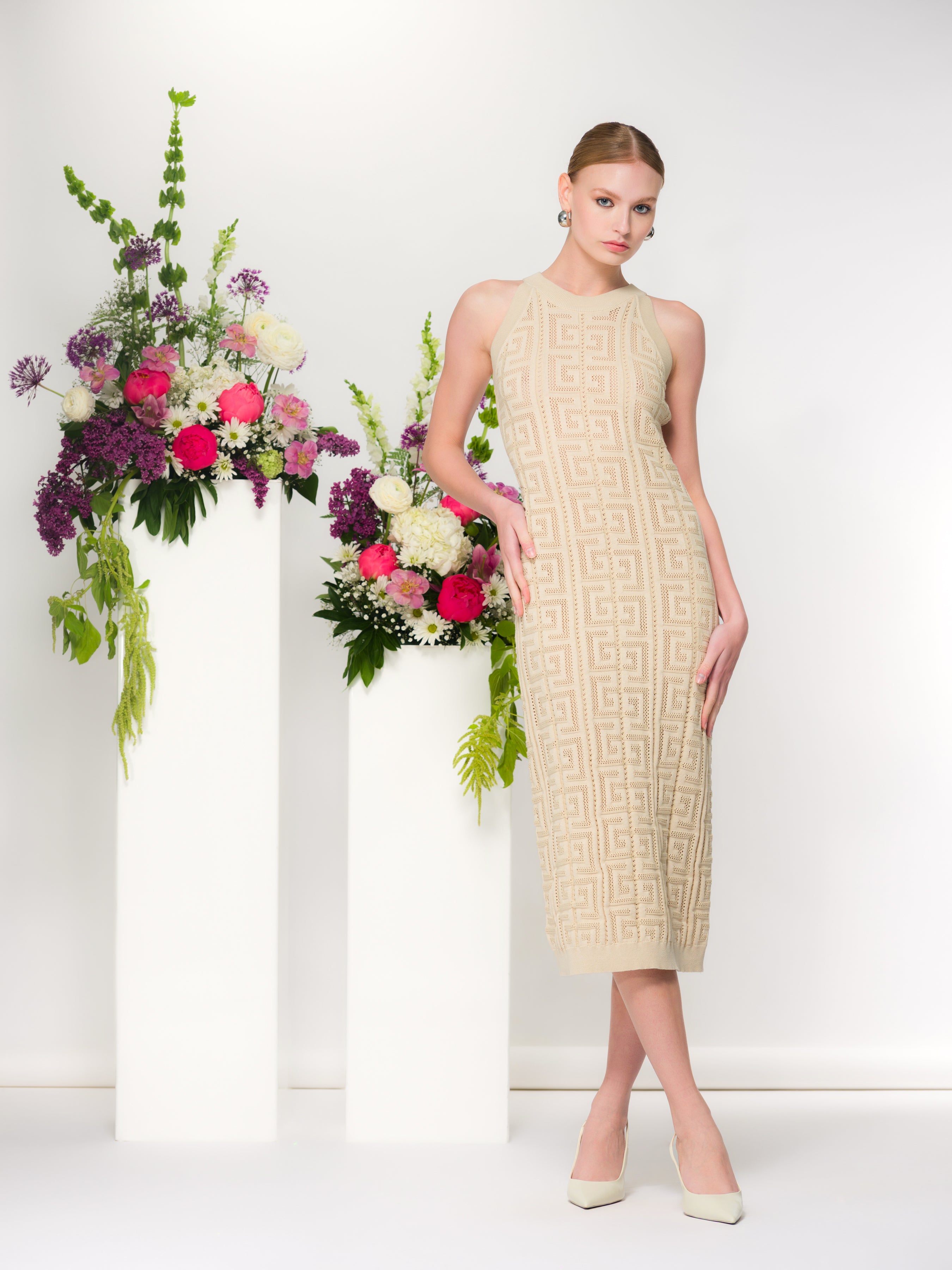 Model wearing a sleeveless textured knit midi dress with a fitted silhouette, styled with pointed-toe heels, posed in a studio setting with floral arrangements on pedestal stands.