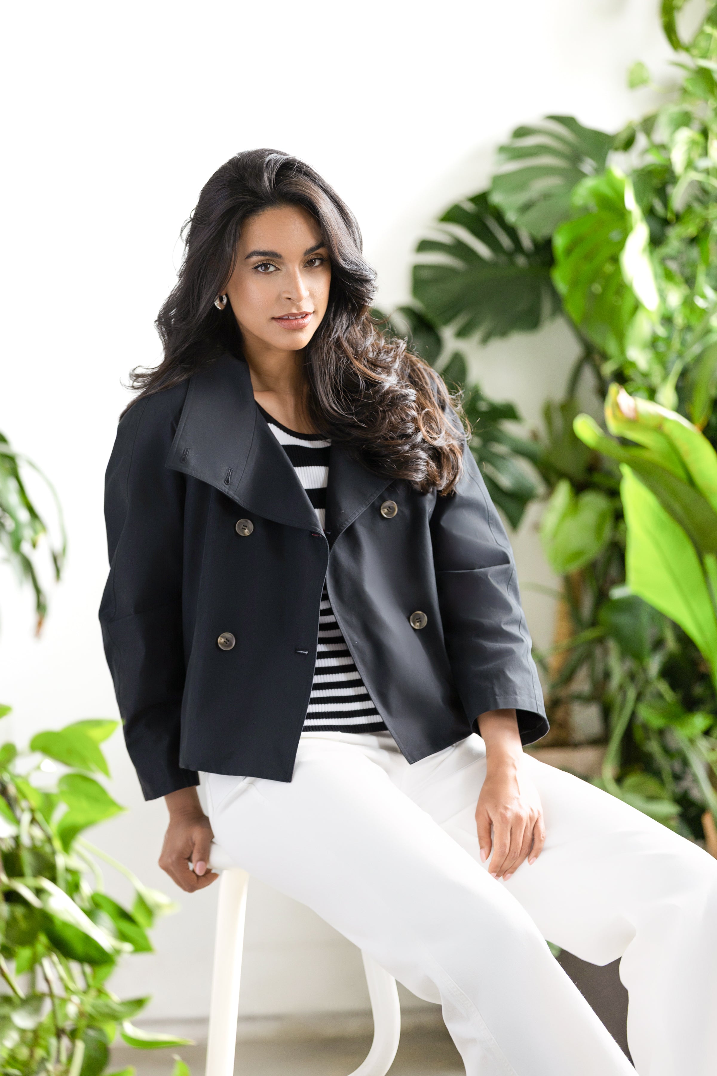 Model wearing a cropped double-breasted jacket with a striped knit top and wide-leg trousers, seated indoors beside lush greenery.