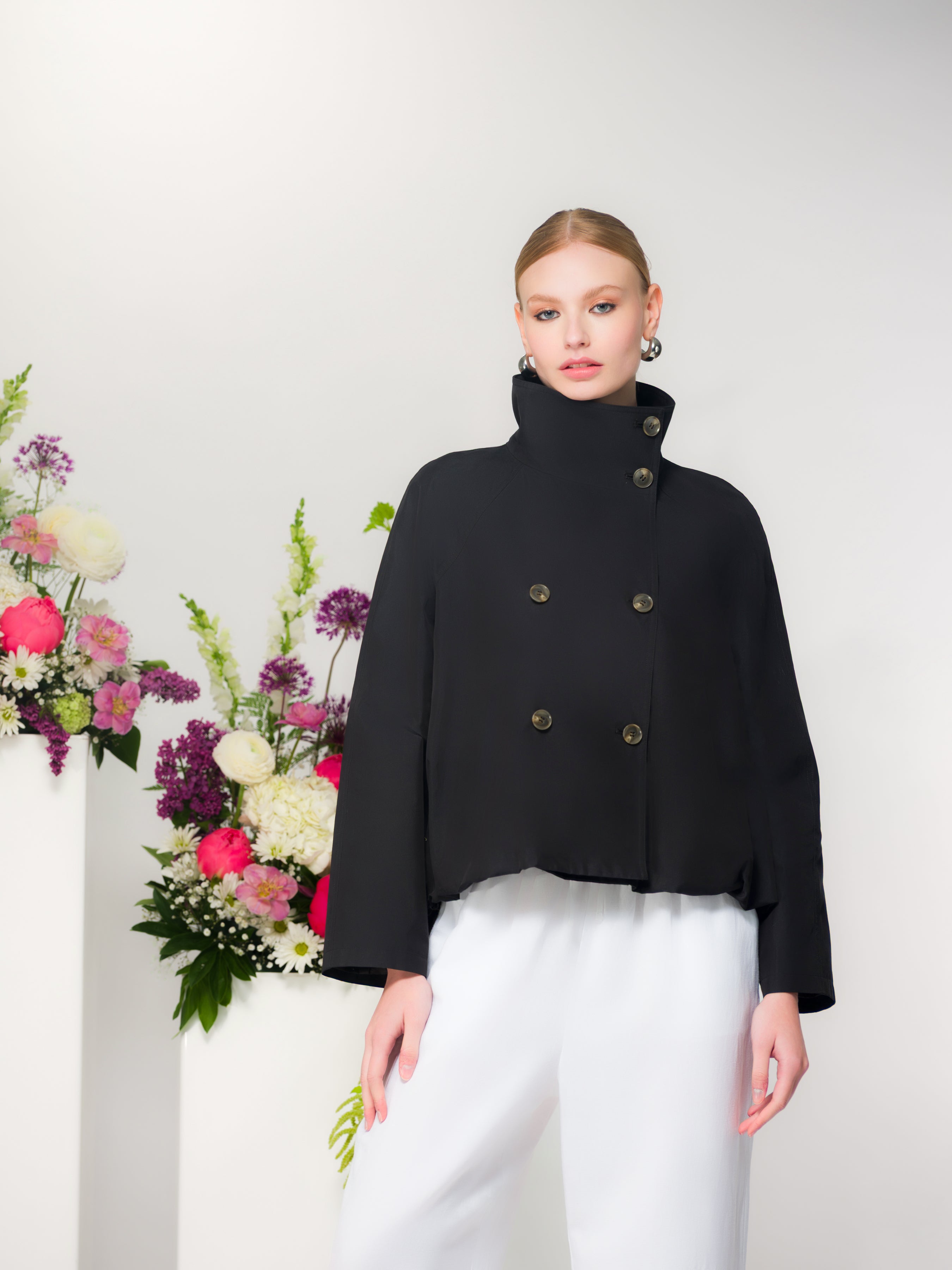 Model wearing a cropped double-breasted jacket with wide-leg trousers, indoors beside floral arrangements.