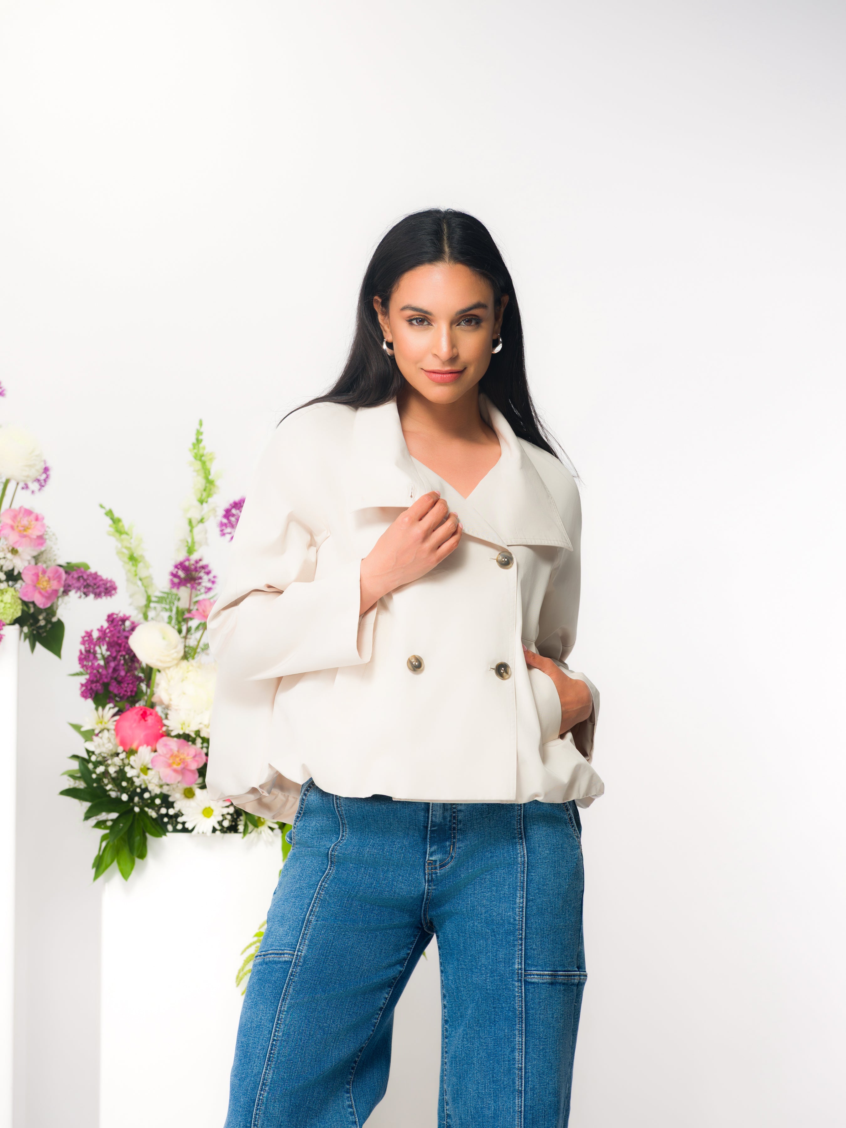Model wearing a cropped double-breasted jacket with jeans, indoors beside floral arrangements.