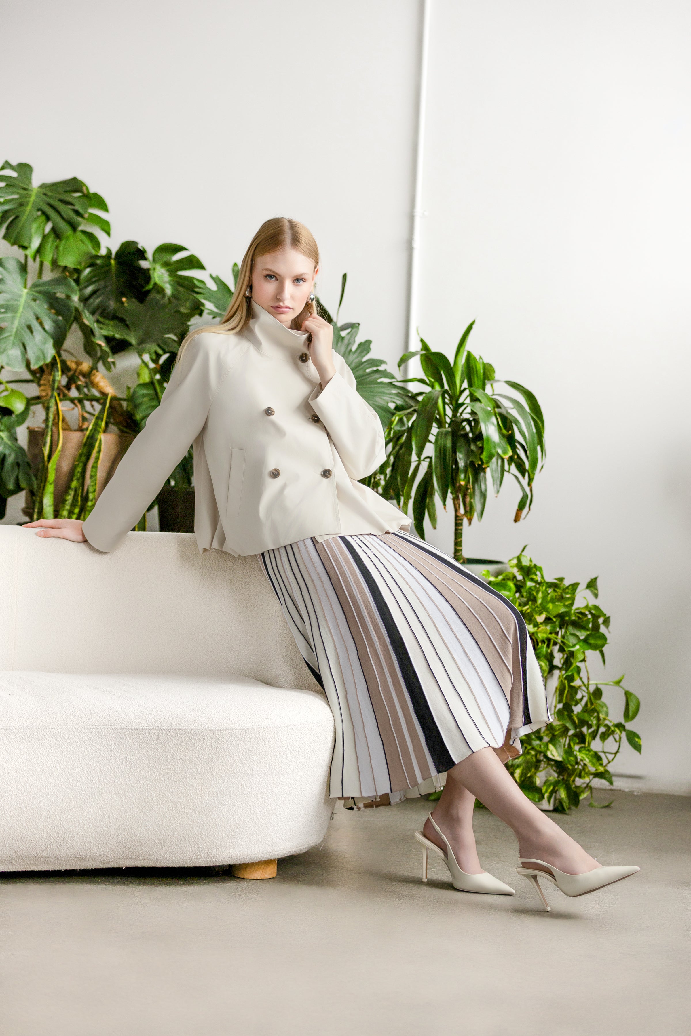 Model wearing a double-breasted jacket styled with a pleated midi skirt and slingback heels, posed on a sofa with indoor plants in the background.