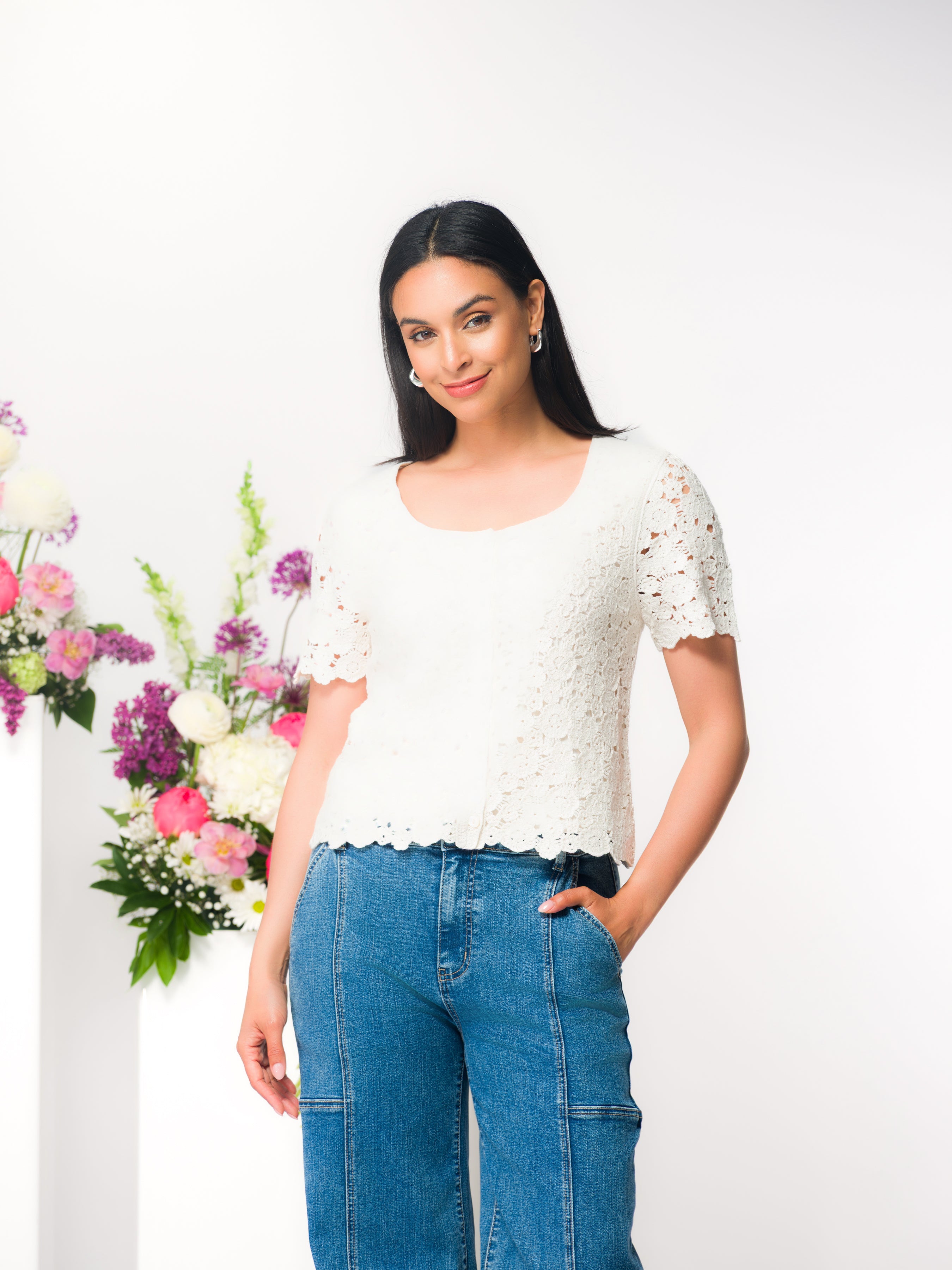 Woman wearing a lace-textured short-sleeve sweater paired with denim pants, posed in a studio with floral accents, showing the sweater’s relaxed fit and delicate knit detail.