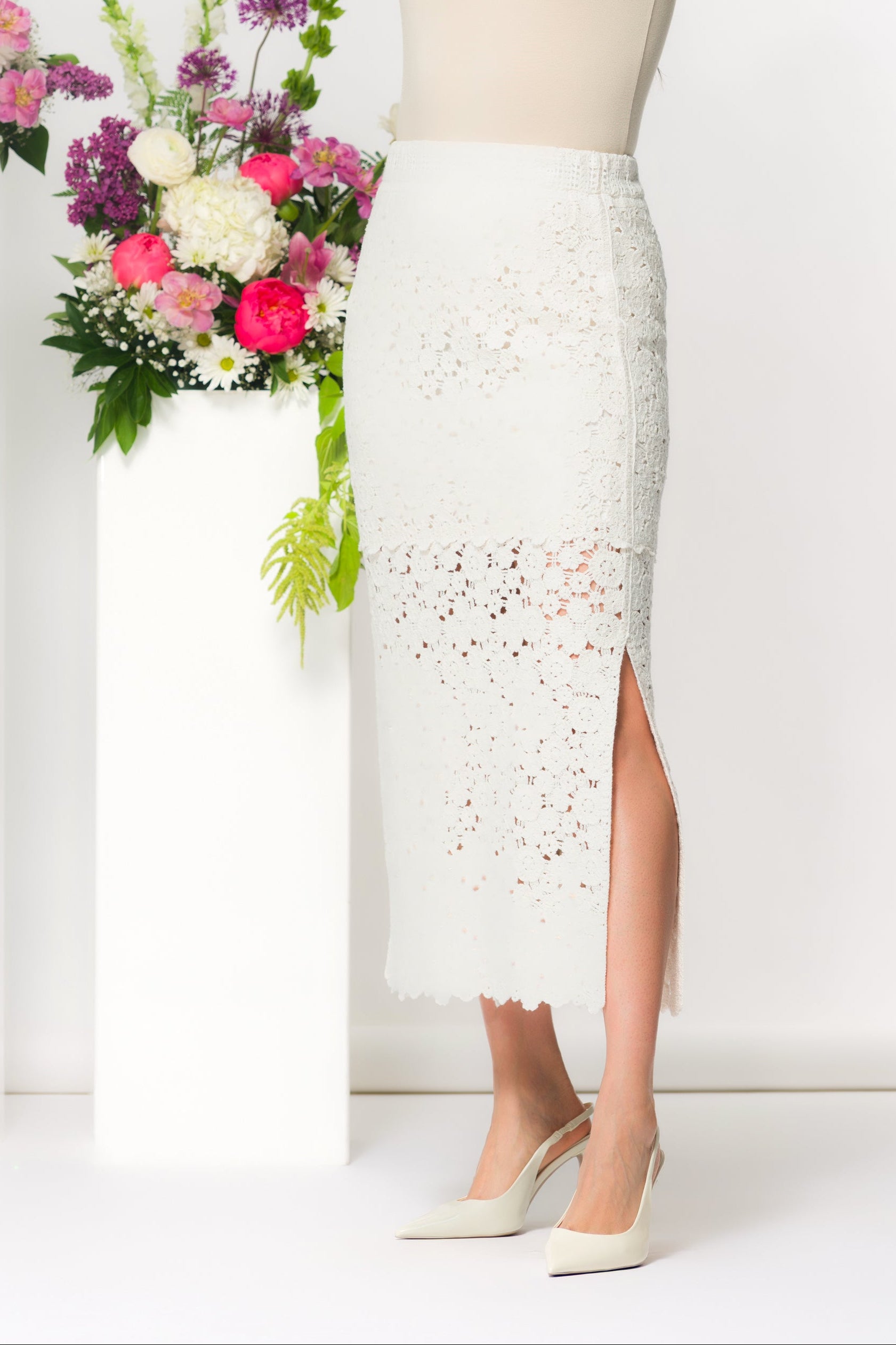 Model wearing a lace knit midi skirt styled with a fitted sleeveless top and pointed slingback heels, posed in a studio setting with floral arrangements.
