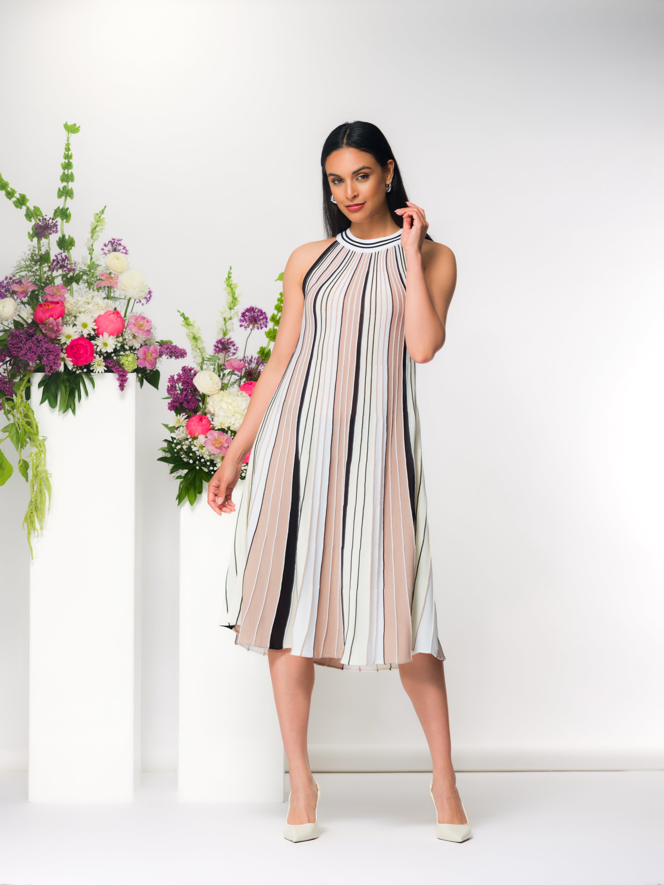 Model in the Hartridge Pleated Knit Dress in beige, cream, and black vertical stripes, showcasing the sleeveless silhouette and fluid pleated midi length in a studio setting with heels.