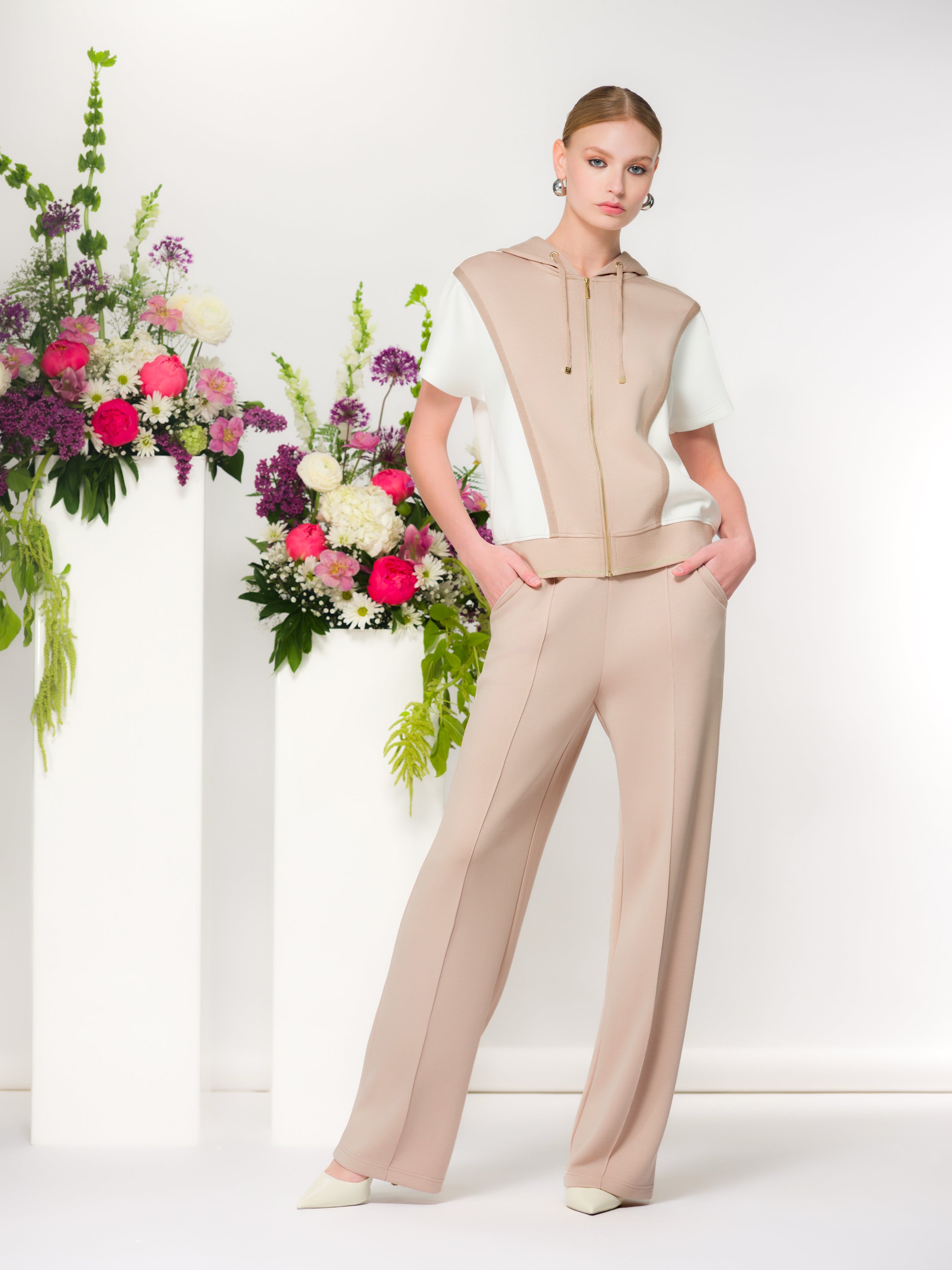 Model wearing a beige and white short-sleeve zip-front hoodie with a hood, paired with sand coloured trousers, styled in a bright indoor setting.