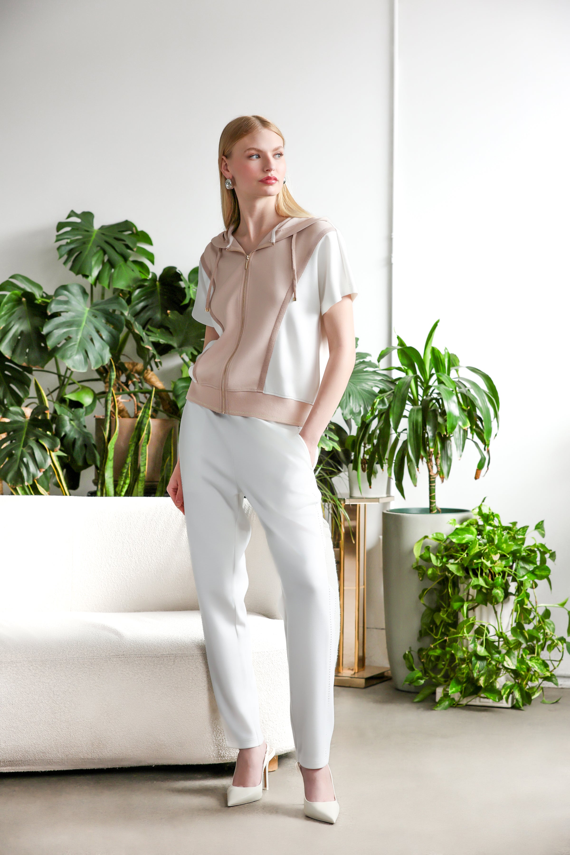 Model wearing a beige and white short-sleeve zip-front hoodie with a hood, paired with light grey slim-leg trousers, styled in a bright indoor setting with green plants.