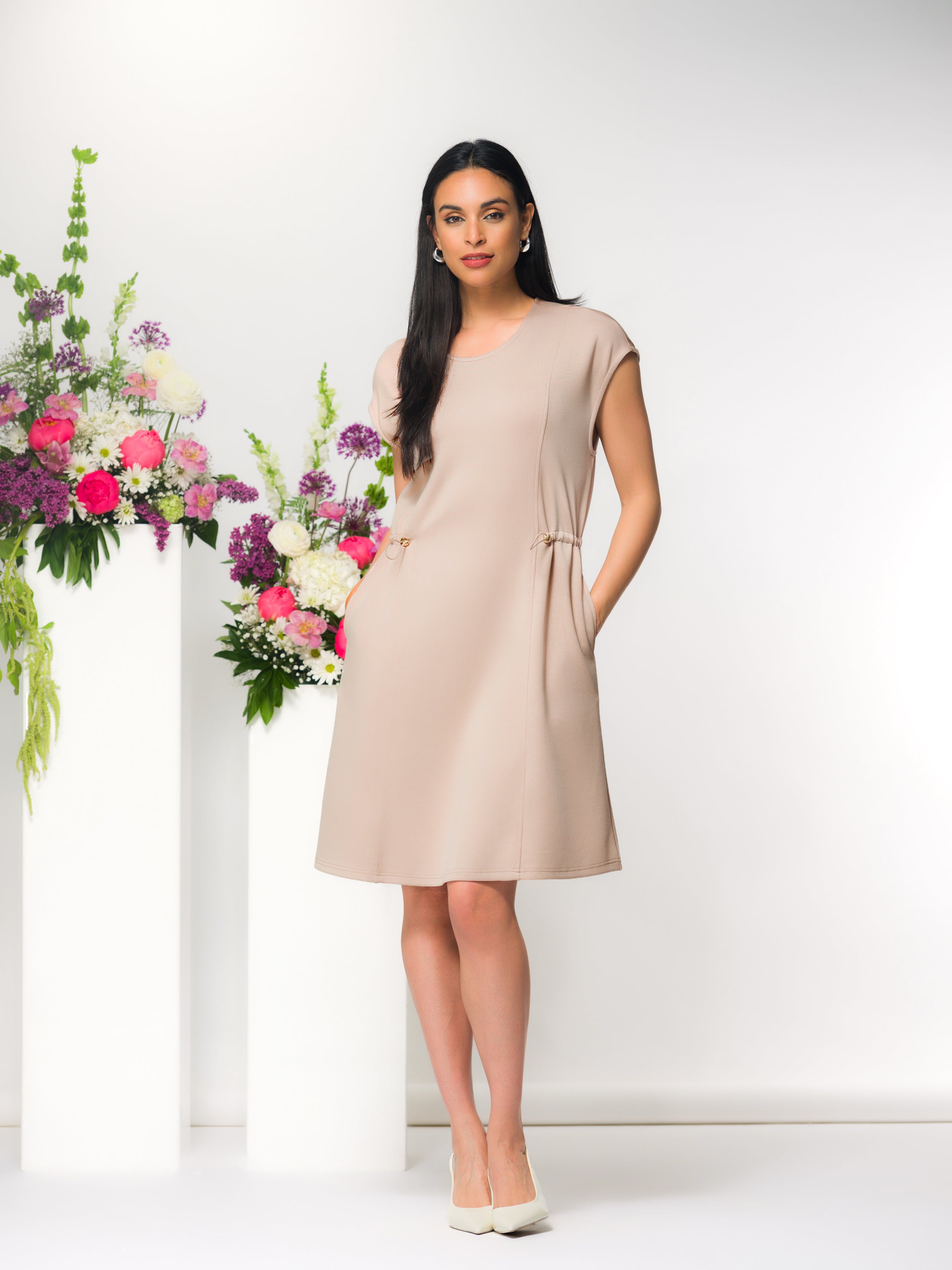 Model wearing a beige knee-length cap sleeve dress with adjustable drawstring waist, side pockets, and round neckline, styled with pointed heels against a studio backdrop with floral arrangements.