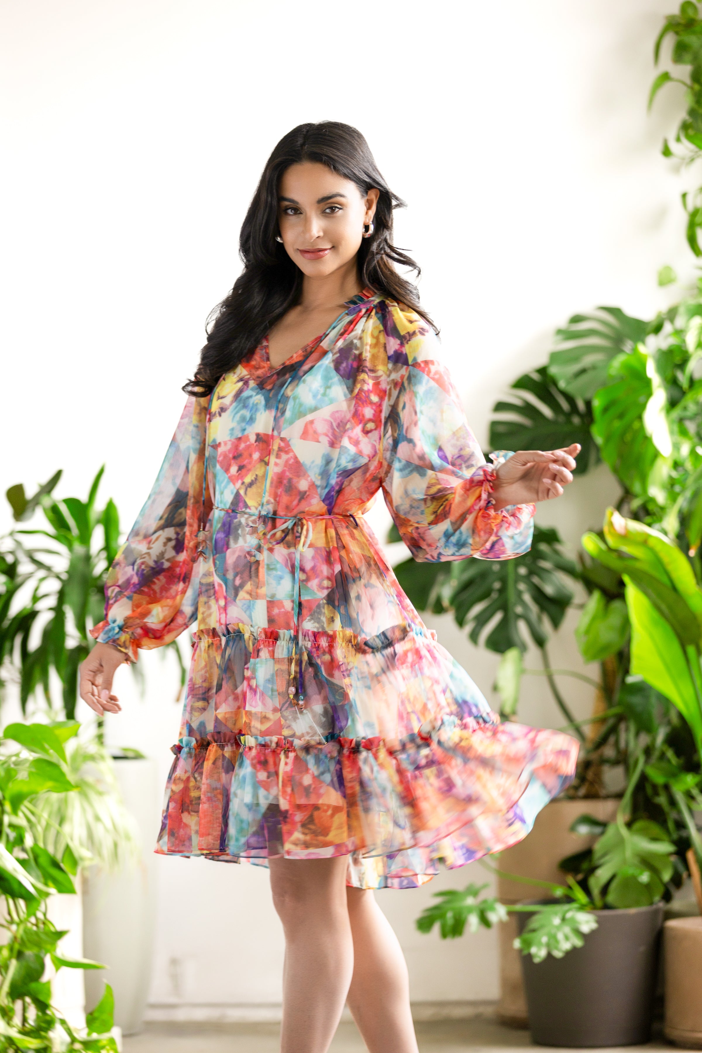 Model wearing a multicolour abstract print chiffon dress with long sheer sleeves and tiered hem, twirling in a bright indoor setting with lush green plants.