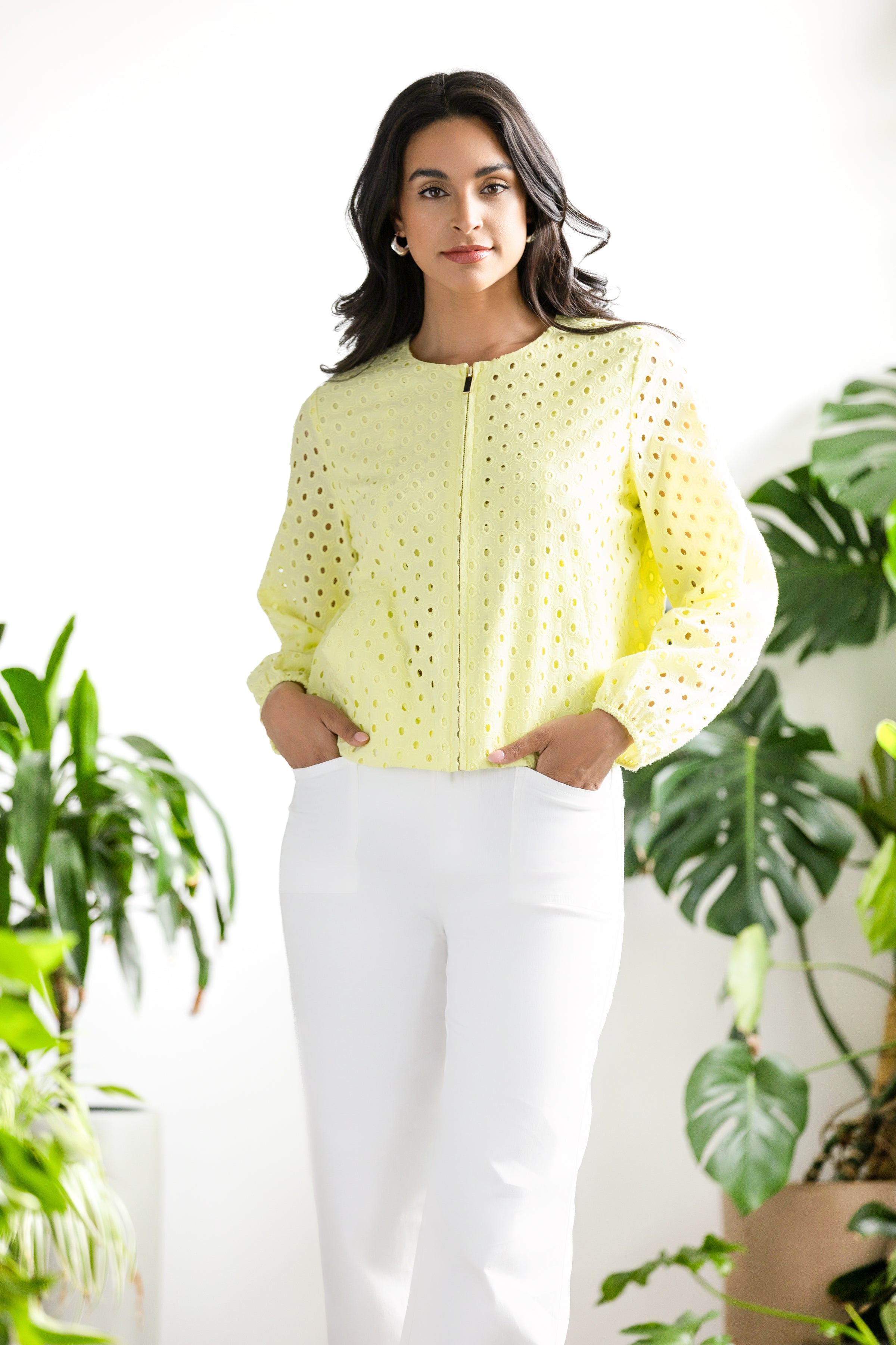 Model wearing a lime green eyelet zip-front cotton jacket with long sleeves and white straight-leg trousers, standing indoors beside green plants.
