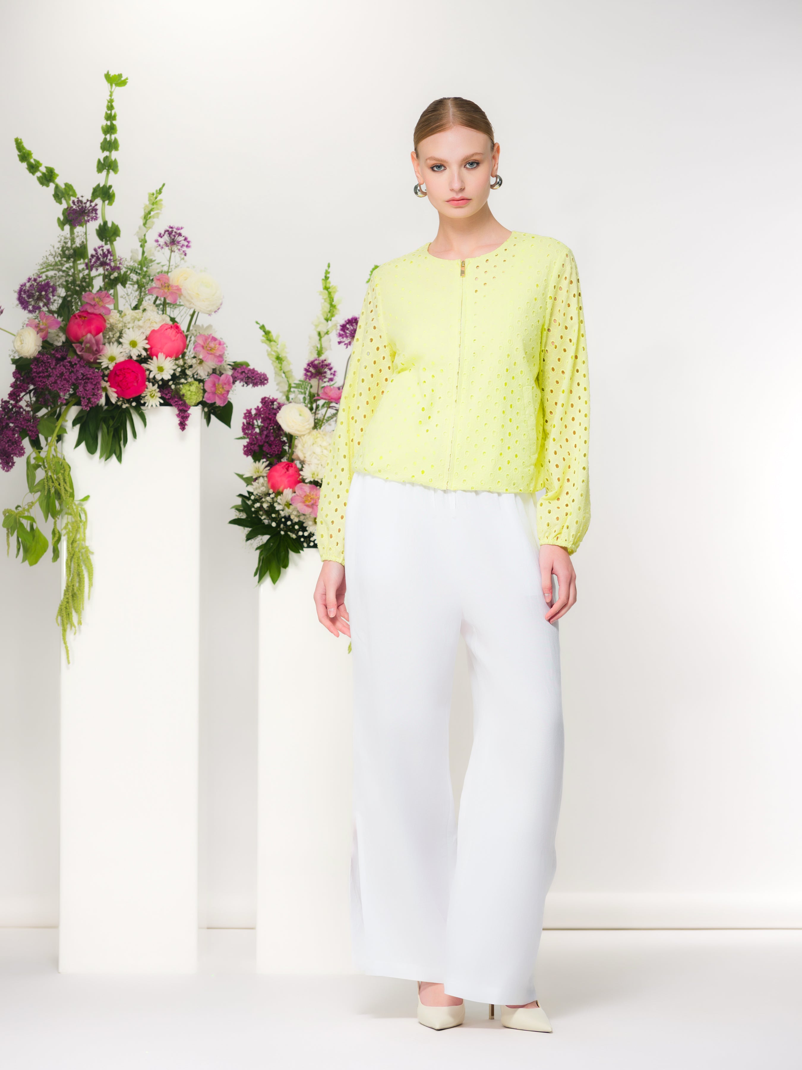 Model wearing a lime green eyelet zip-front cotton jacket with long sleeves and white straight-leg trousers, standing indoors beside floral arrangements.