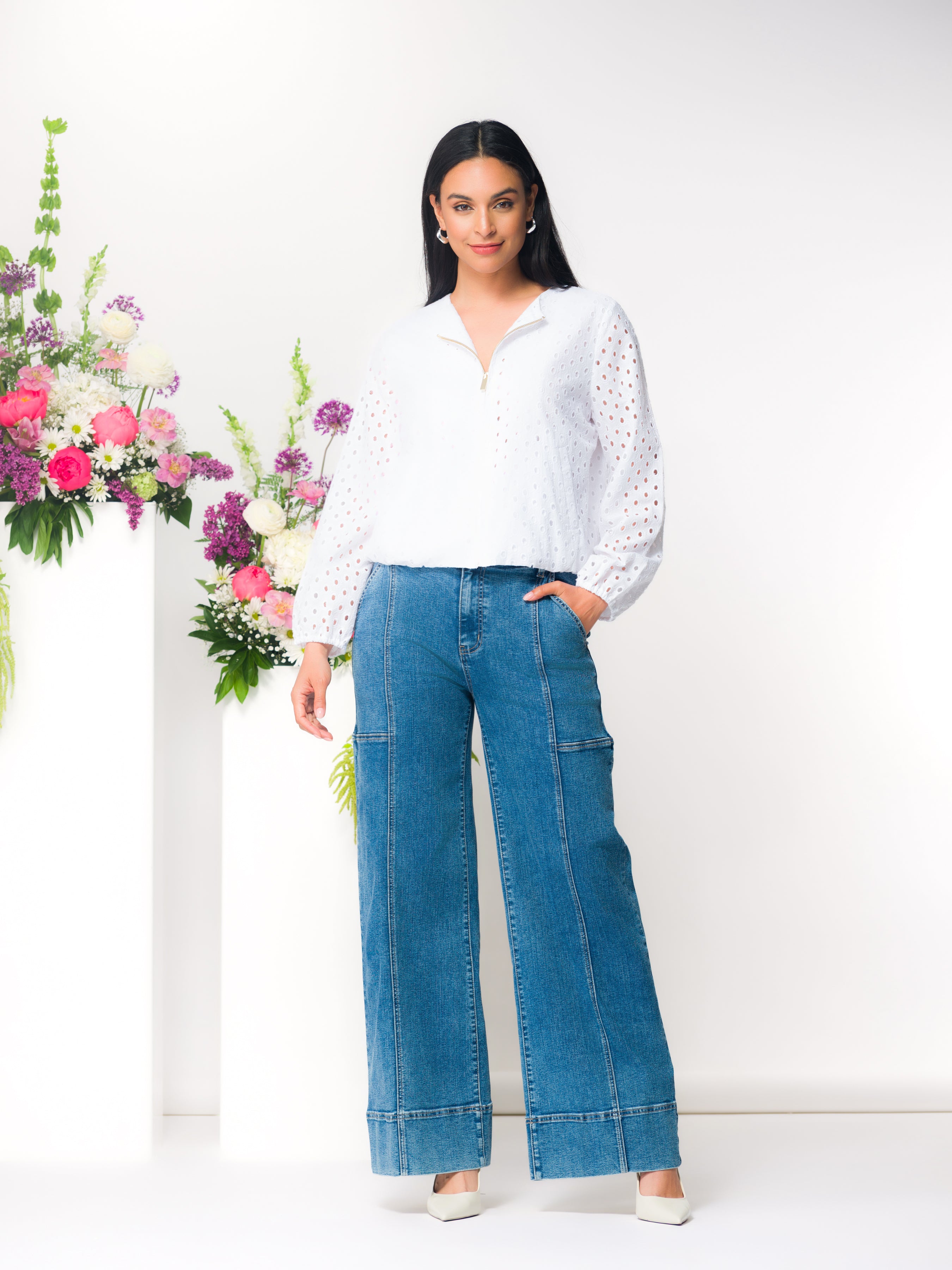 Model wearing a white eyelet blouse with a half-zip neckline and long sleeves, paired with wide-leg blue jeans and neutral heels, standing beside a floral arrangement.