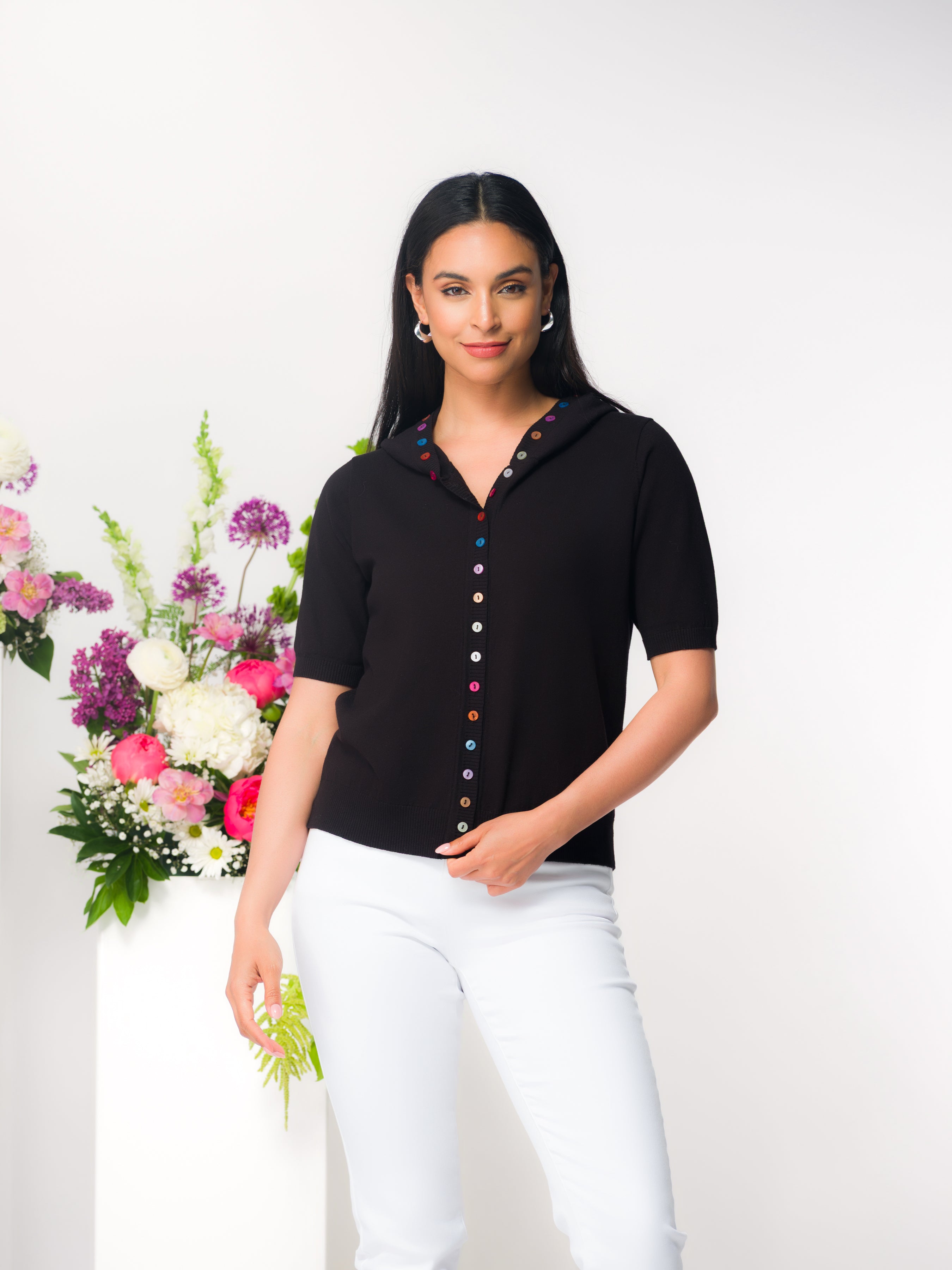 Model wearing a black short-sleeve hooded cardigan with multicolour button-front detail, paired with white pants.