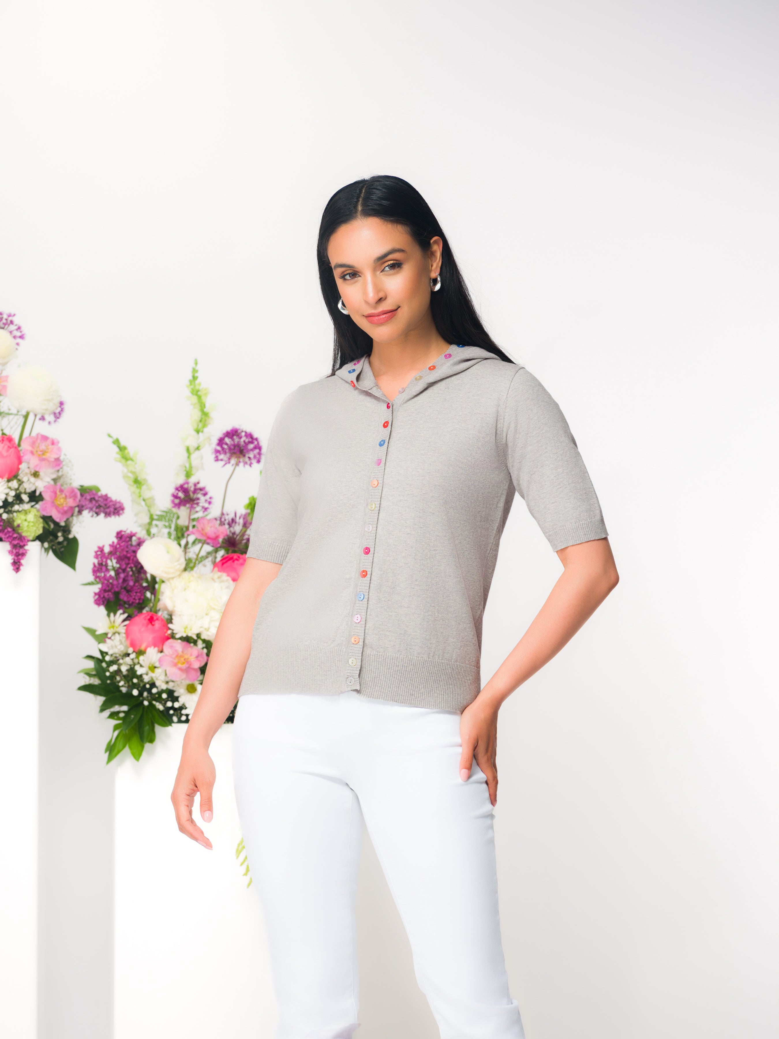 Model wearing a light grey short-sleeve hooded cardigan with multicolour buttons, styled with white pants.