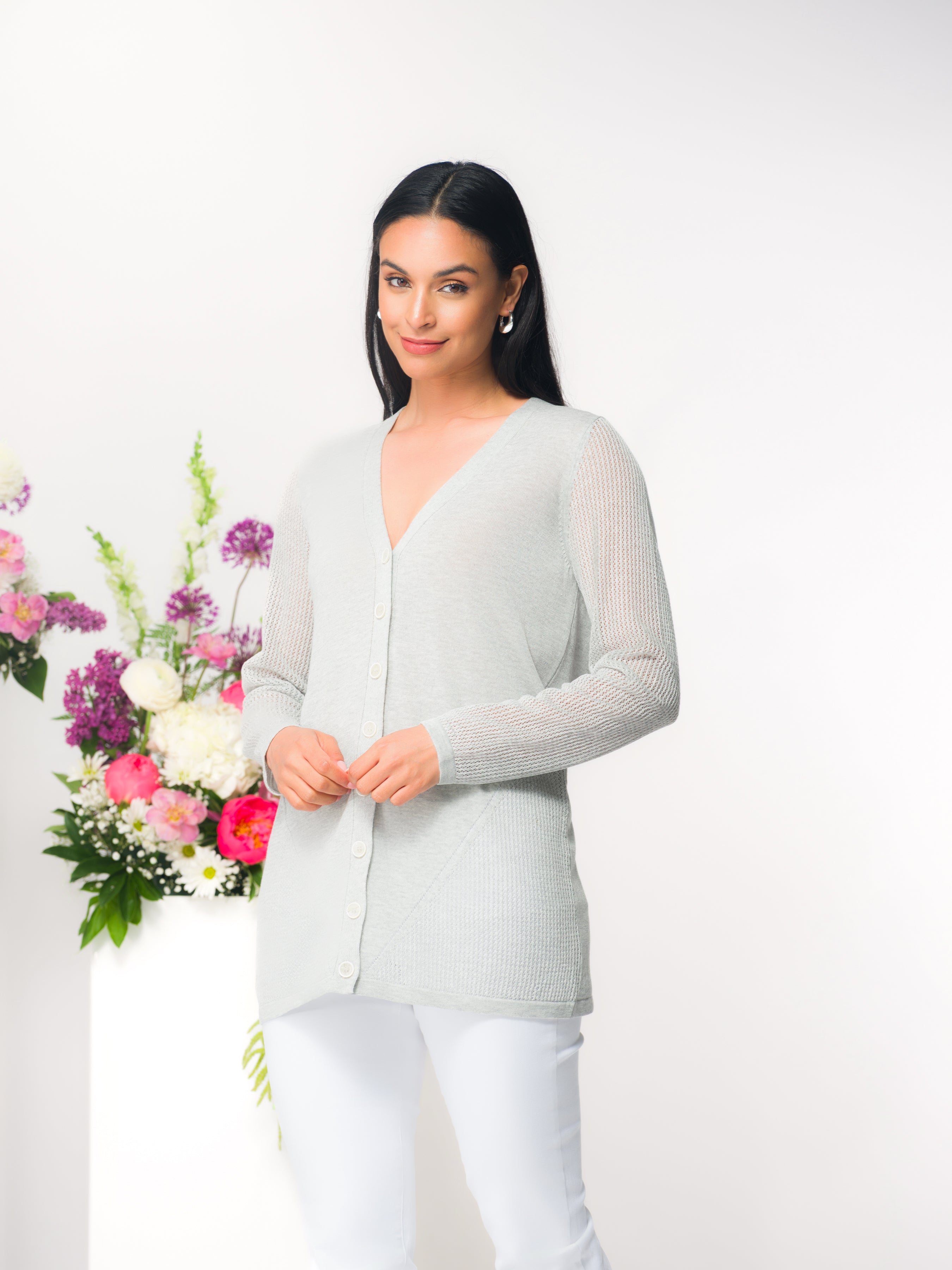 Full-length image of a model wearing a light grey V-neck button-front cardigan with long sleeves and subtle textured knit detailing, paired with slim white pants. She stands against a clean white backdrop beside a pedestal arrangement of pink, white, and purple flowers.
