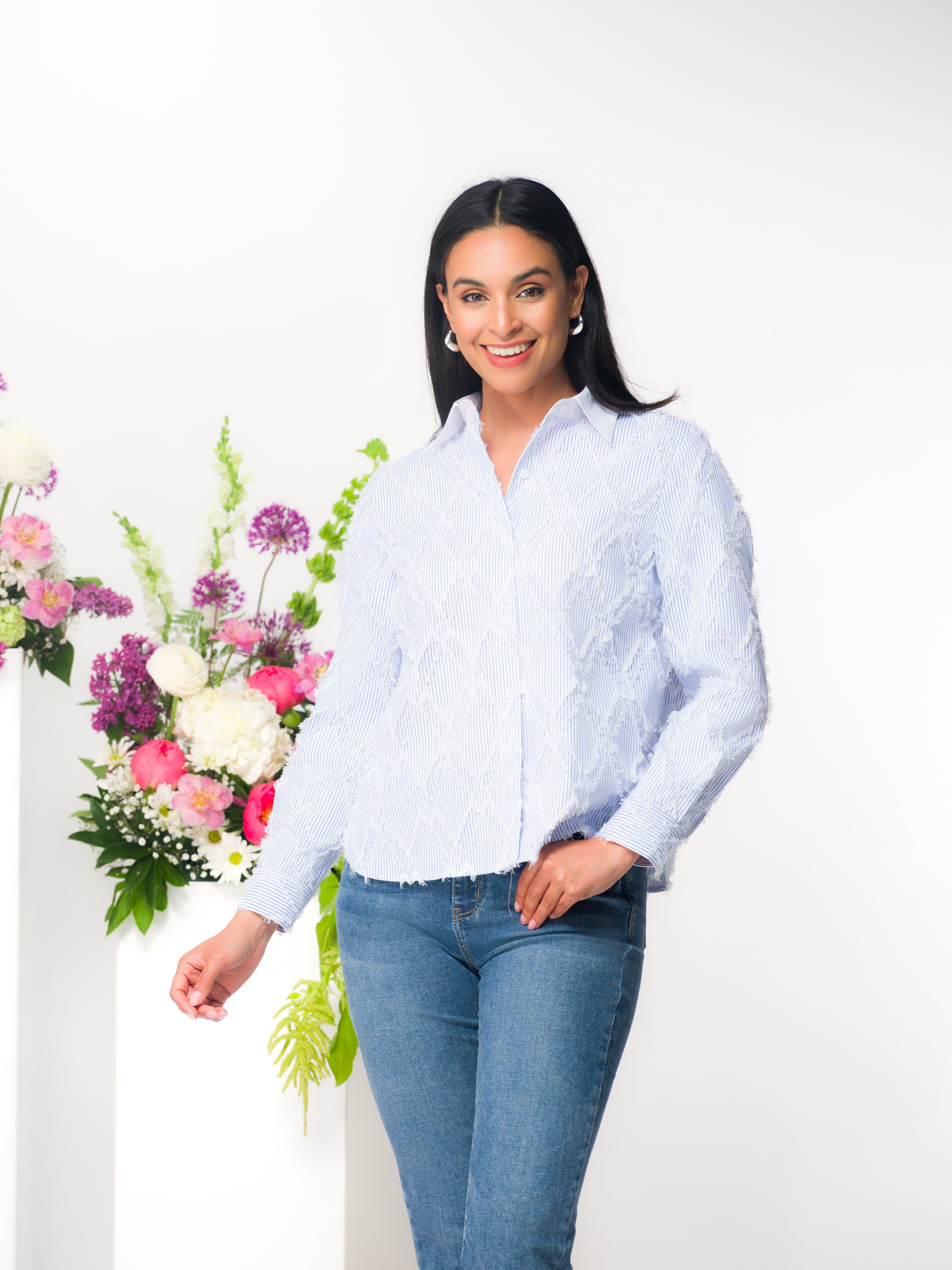 Smiling woman modelling a light blue textured button-front blouse with long sleeves and a collar, styled with fitted medium-wash jeans, standing beside a floral arrangement against a neutral studio background.