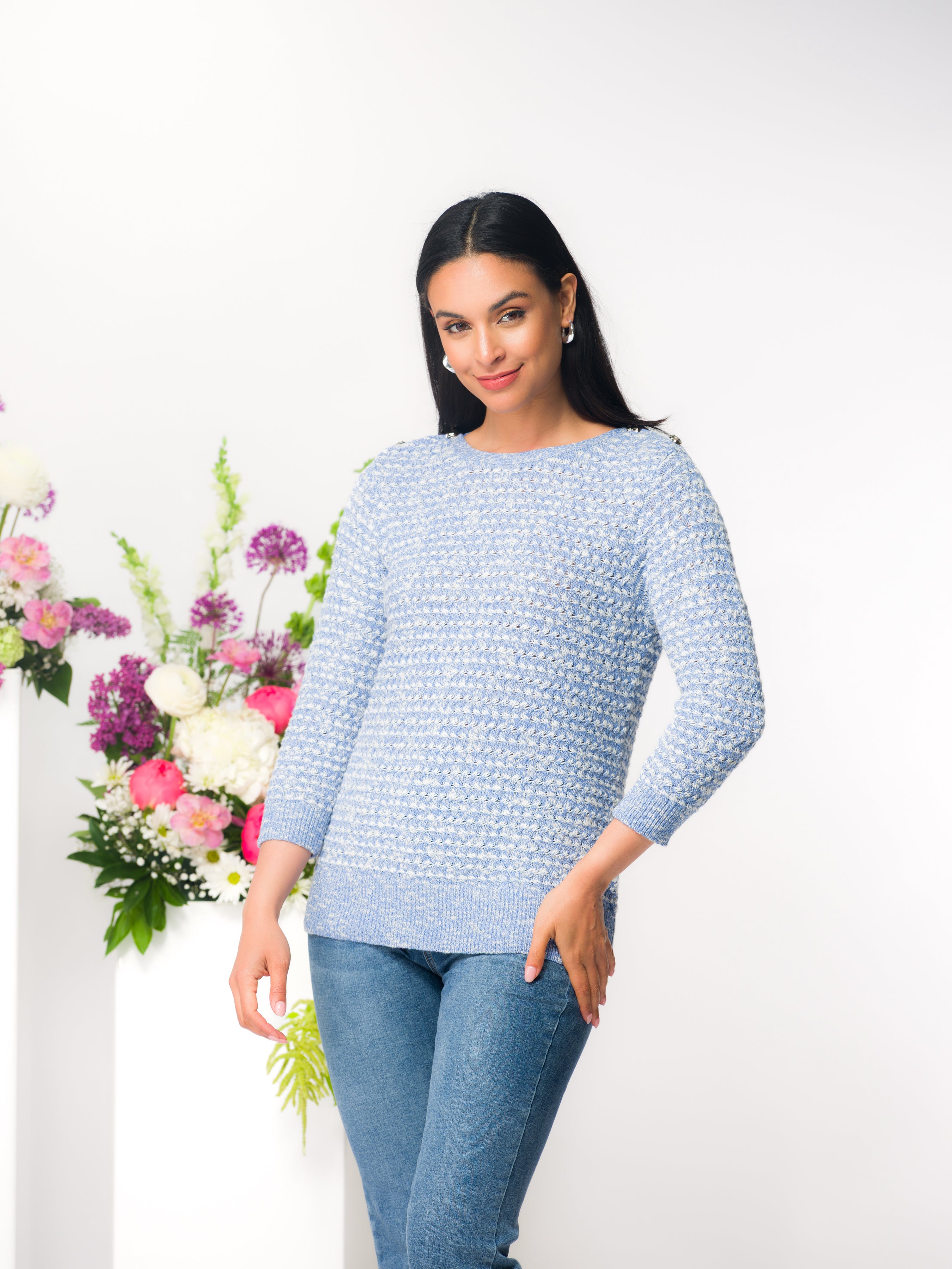 Woman wearing a light blue textured cotton sweater with a classic crew neckline and three-quarter sleeves, paired with medium-wash jeans. She stands in a bright studio setting beside a floral arrangement, modelling the relaxed fit and soft knit texture of the sweater.