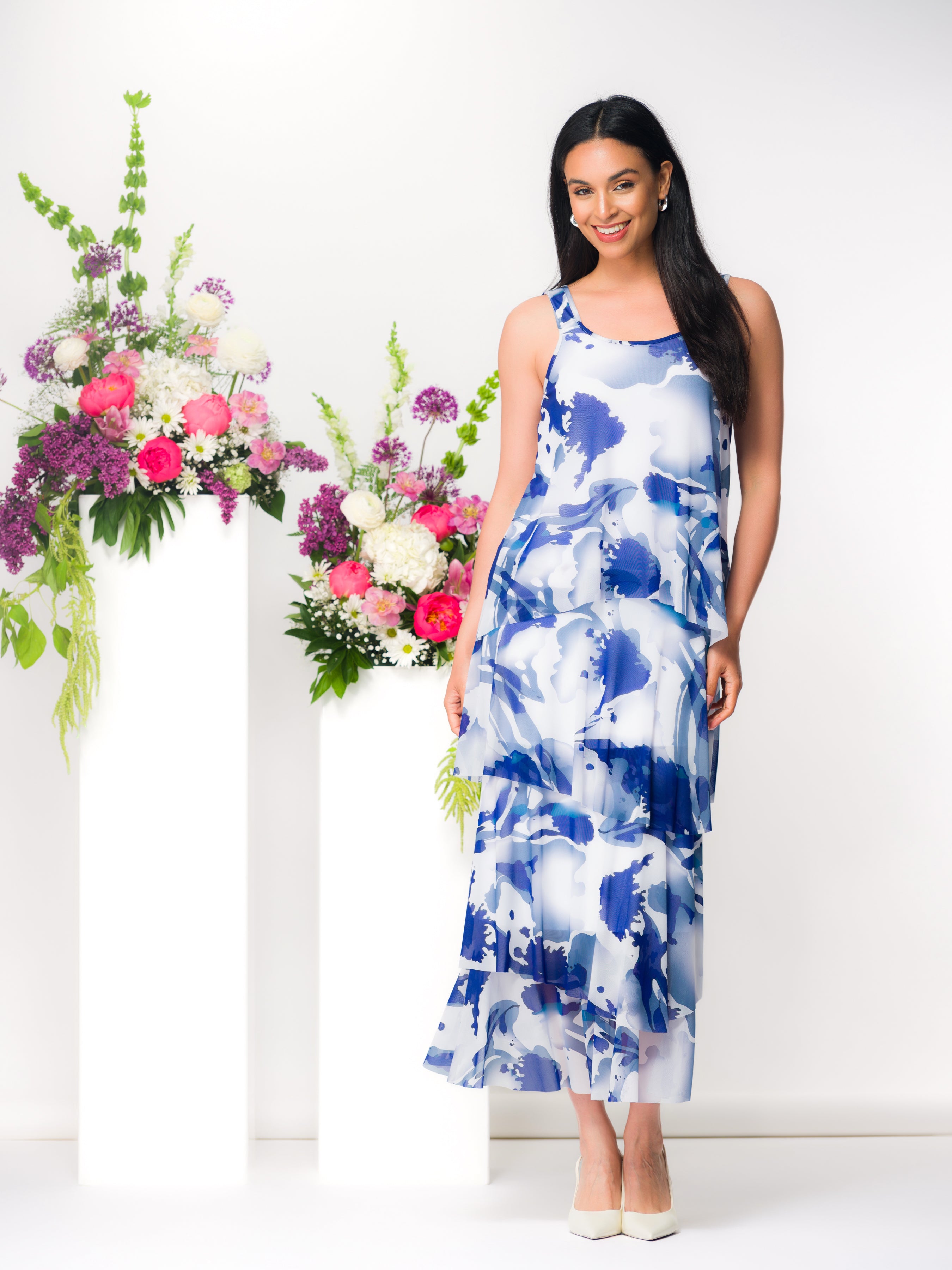 Smiling woman wearing a blue and white abstract floral print sleeveless midi dress with a tiered, flowy skirt, paired with ivory pointed-toe heels, standing beside white pedestals topped with pink and white floral arrangements against a light grey background.