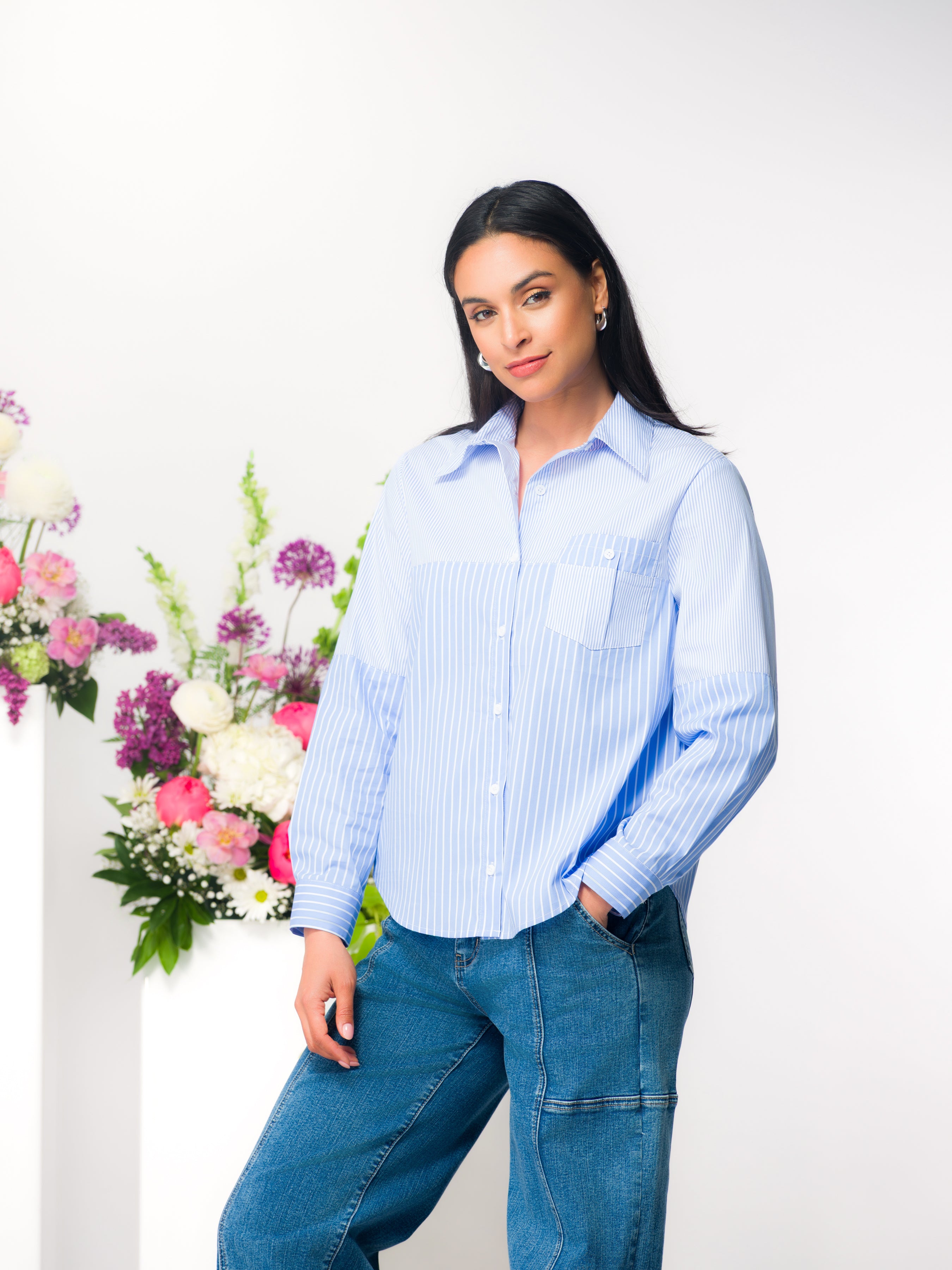 Model wearing a light blue vertical stripe collared blouse with chest pocket, paired with medium wash jeans, standing beside a floral arrangement in a studio setting.