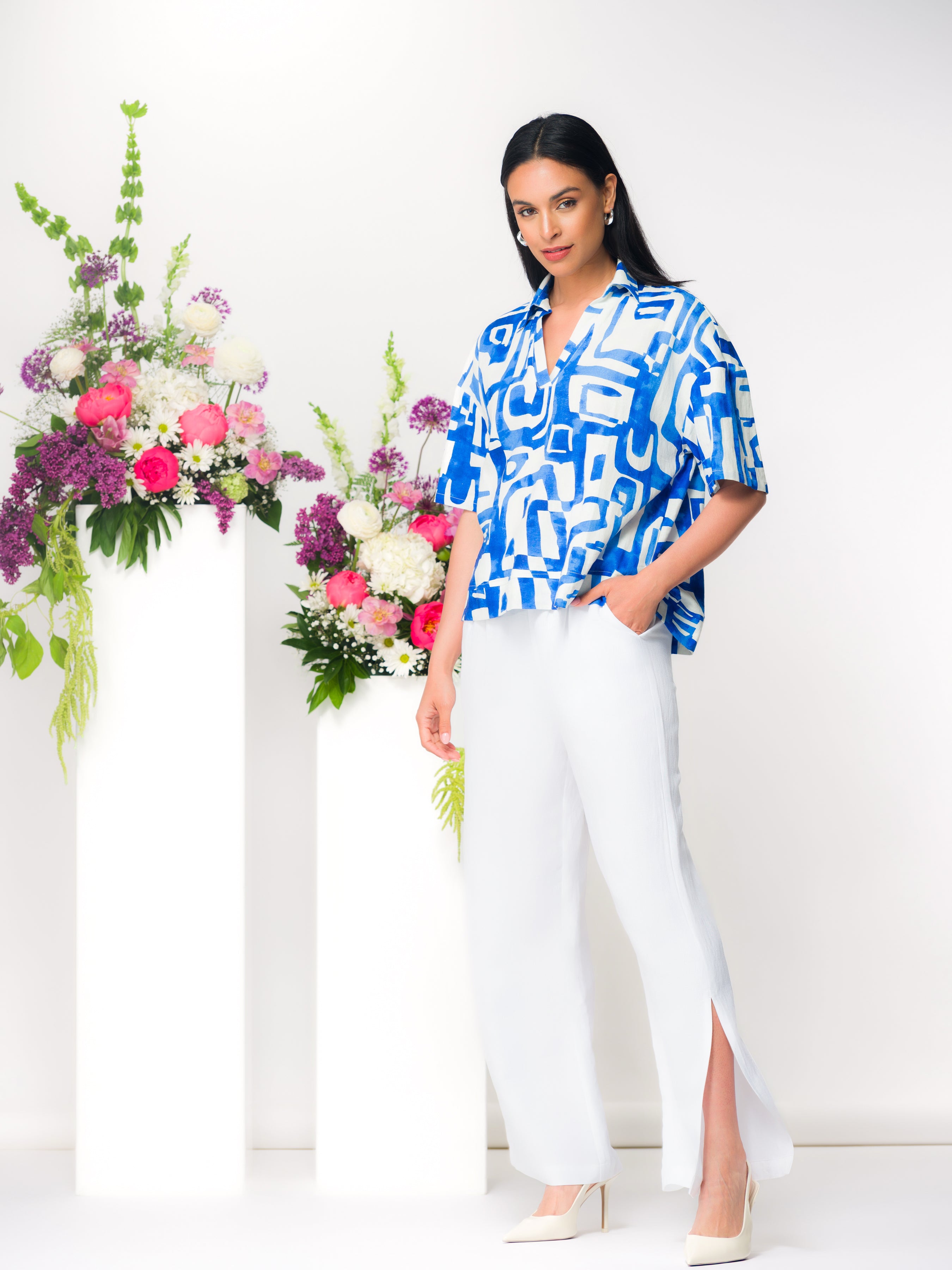 Model wearing a blue and white abstract print blouse with light grey wide-leg trousers and cream heels, posed beside floral arrangements in a bright studio.