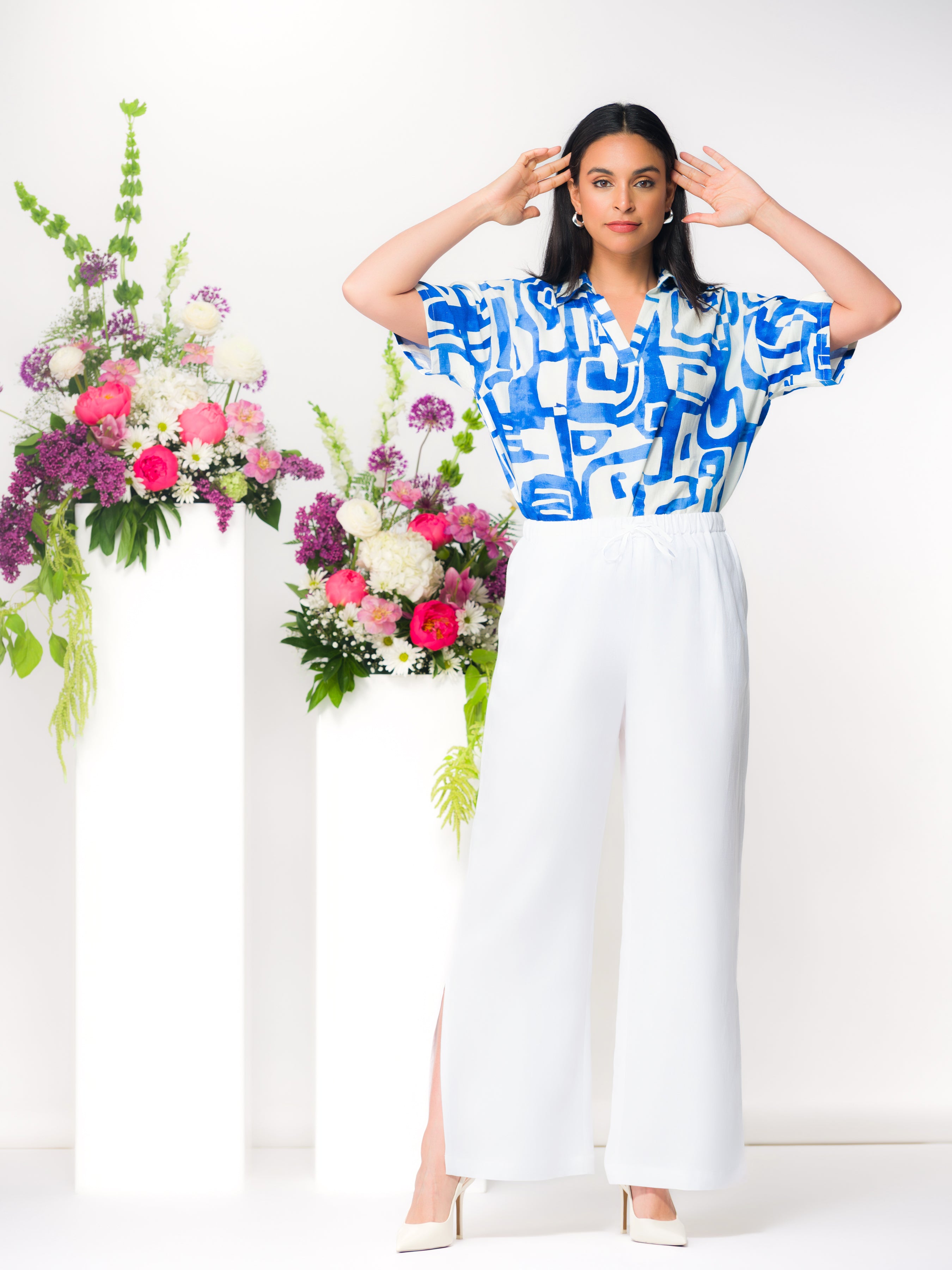 Model wearing white wide-leg linen blend pants with a drawstring waist and side slit, styled with a patterned blouse and heels in a bright studio setting.