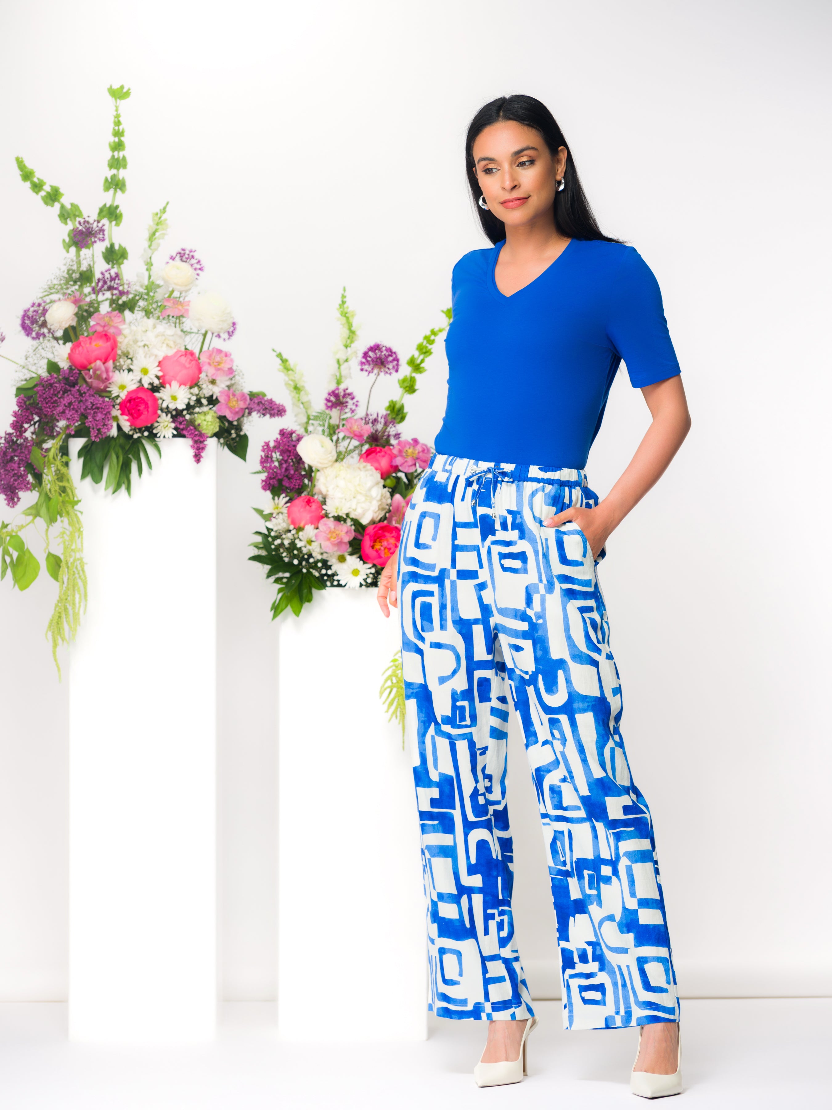 Model wearing a cobalt V-neck top with blue and white geometric wide-leg drawstring linen-blend pants and neutral heels, standing beside floral displays in a bright studio.