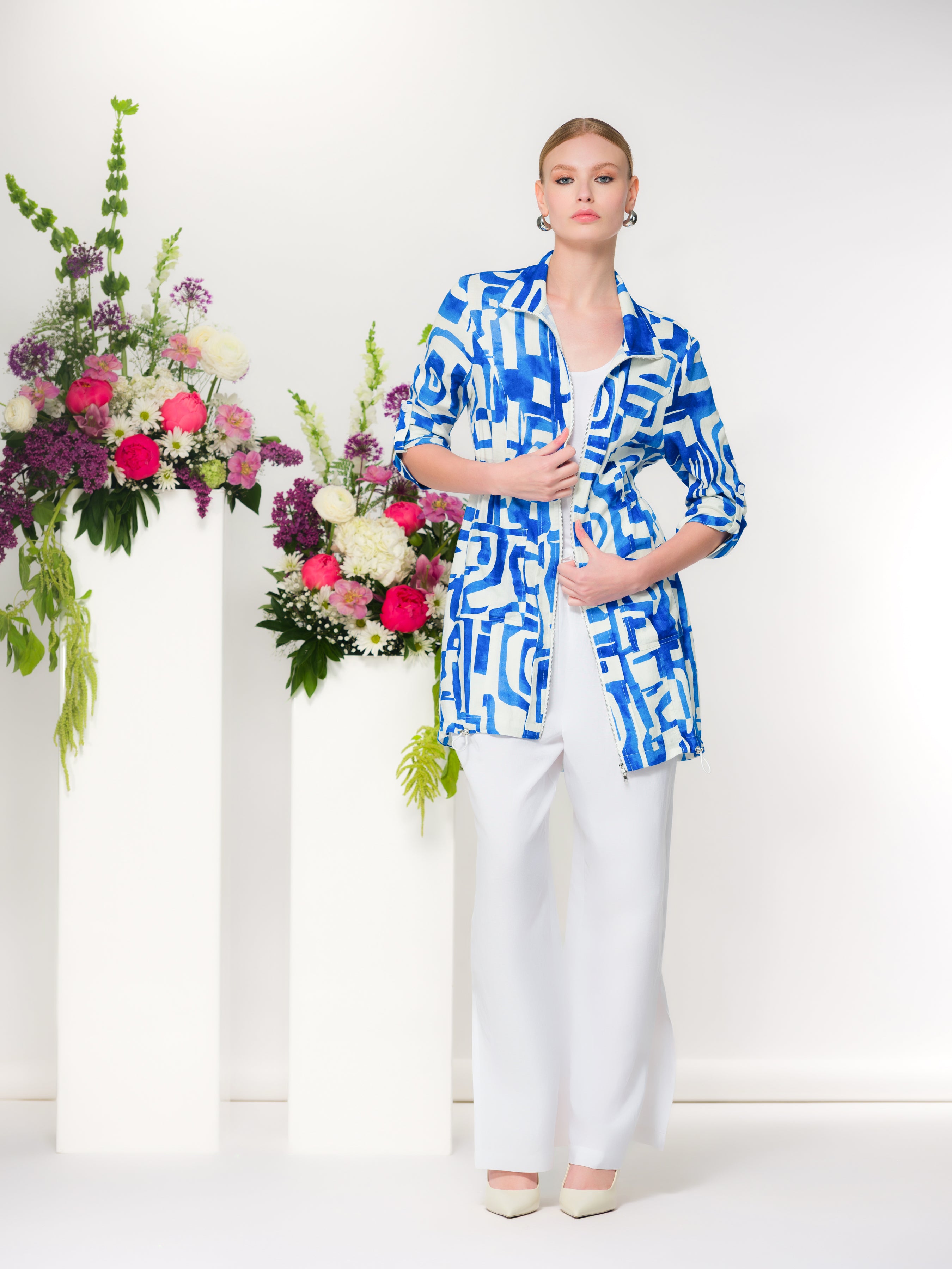 Model wearing a blue and white abstract-print linen blend zip-front jacket with a stand collar and adjustable drawstring waist, styled over a white top and wide-leg white trousers with pointed heels against a minimalist backdrop with floral arrangements.