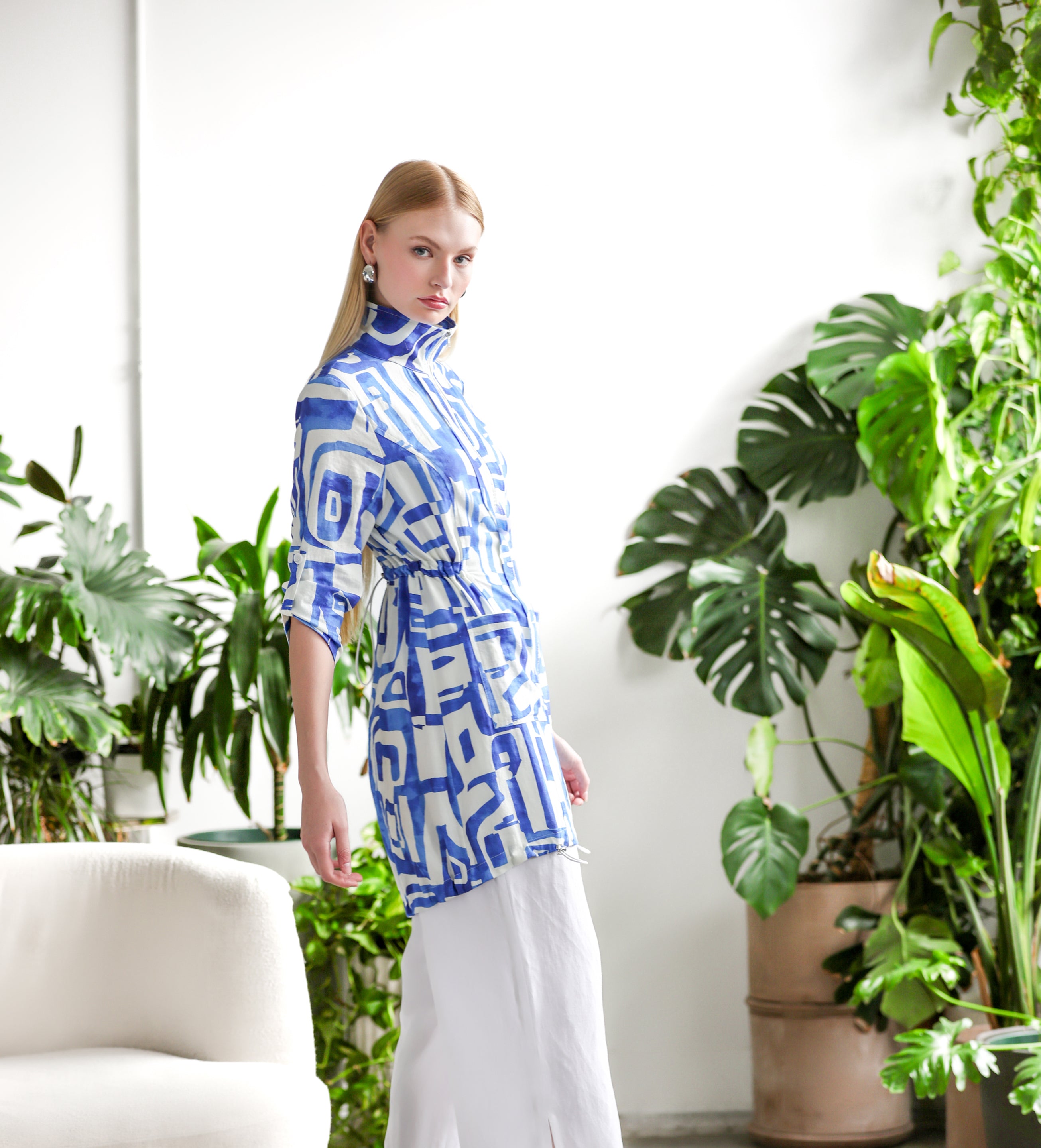 Side view of the blue and white abstract-print tunic-length jacket featuring a zip-front closure, stand collar, and cinched waist detail, paired with flowy white wide-leg pants and pointed heels in a bright room with lush green plants.