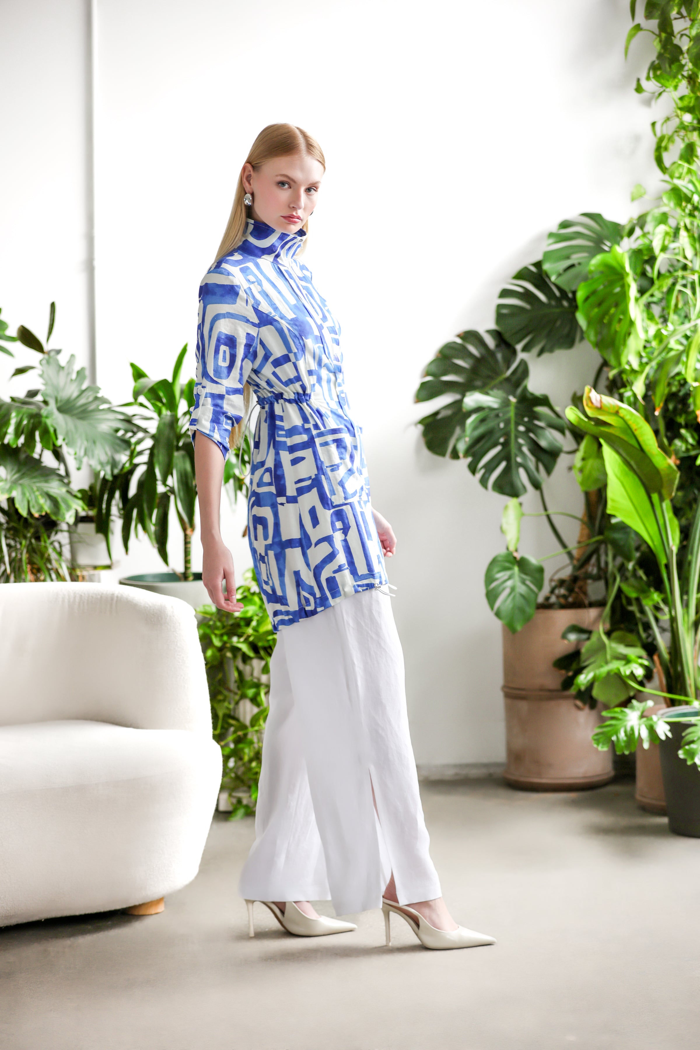 Side view of the blue and white abstract-print linen blend jacket highlighting the adjustable waist and lightweight drape, styled with white wide-leg trousers and pointed heels in a modern indoor setting with greenery.
