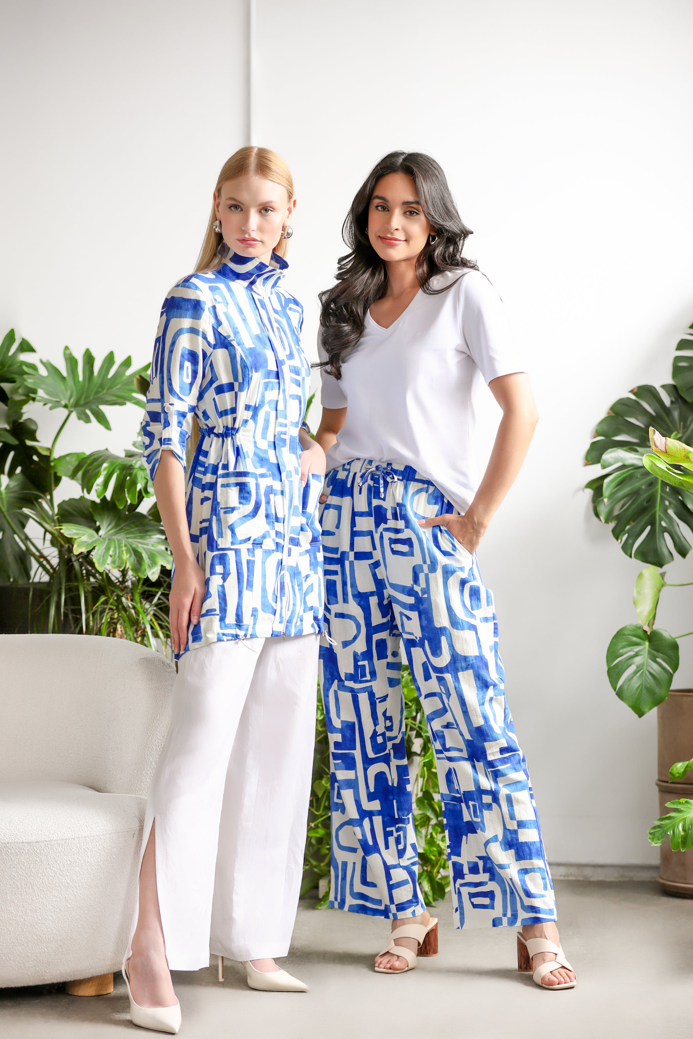 Two women posing indoors beside large green houseplants and a cream-colored chair. One wears a blue-and-white geometric patterned tunic with white wide-leg pants and pointed heels. The other wears a white V-neck top with blue-and-white patterned wide-leg pants and beige heeled sandals