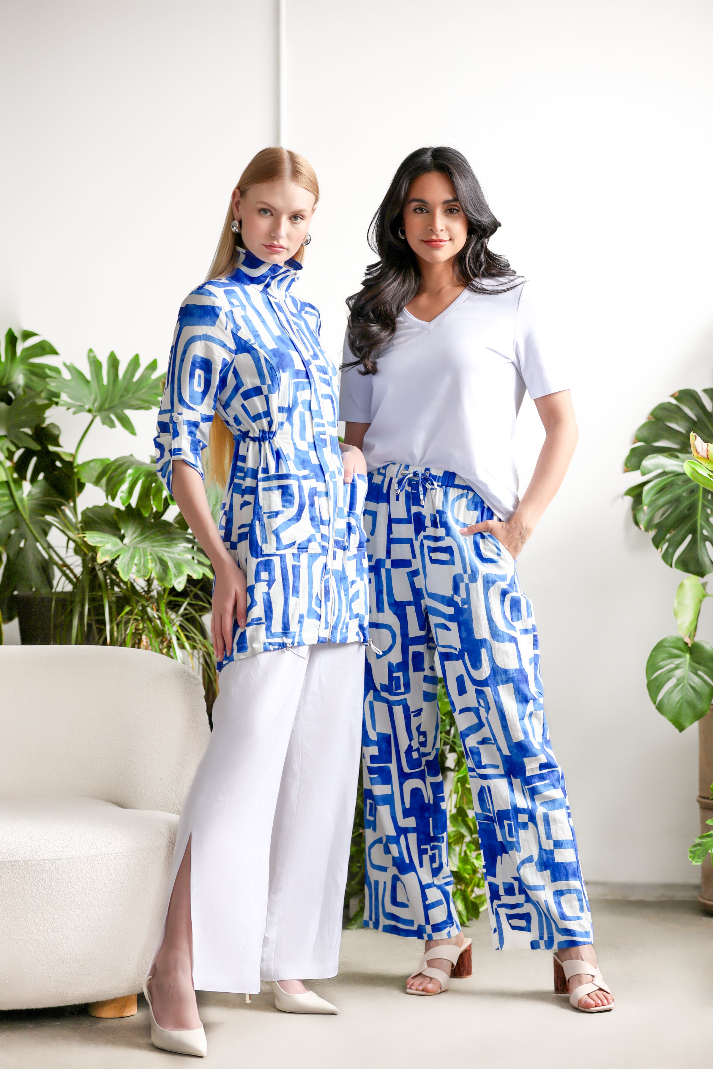 Two women standing together in a bright room with large leafy plants. One is dressed in a blue-and-white abstract print tunic with white wide-leg pants and heels. The other wears a white V-neck shirt paired with matching blue-and-white patterned wide-leg pants and beige heeled sandals.