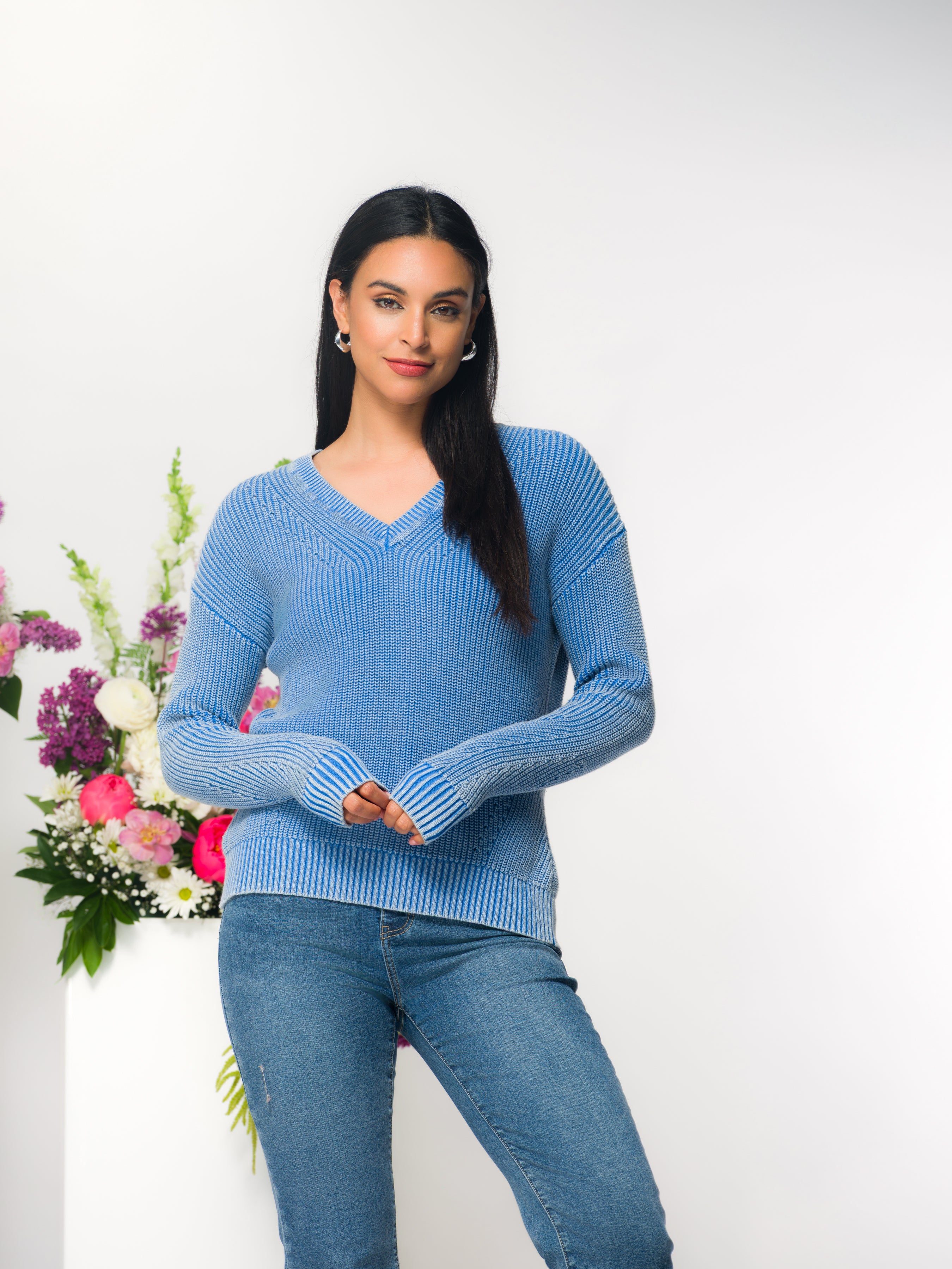 Woman wearing a blue textured V-neck cotton sweater with long sleeves and ribbed cuffs, paired with fitted blue jeans, standing in a studio setting with a neutral backdrop and floral arrangement.