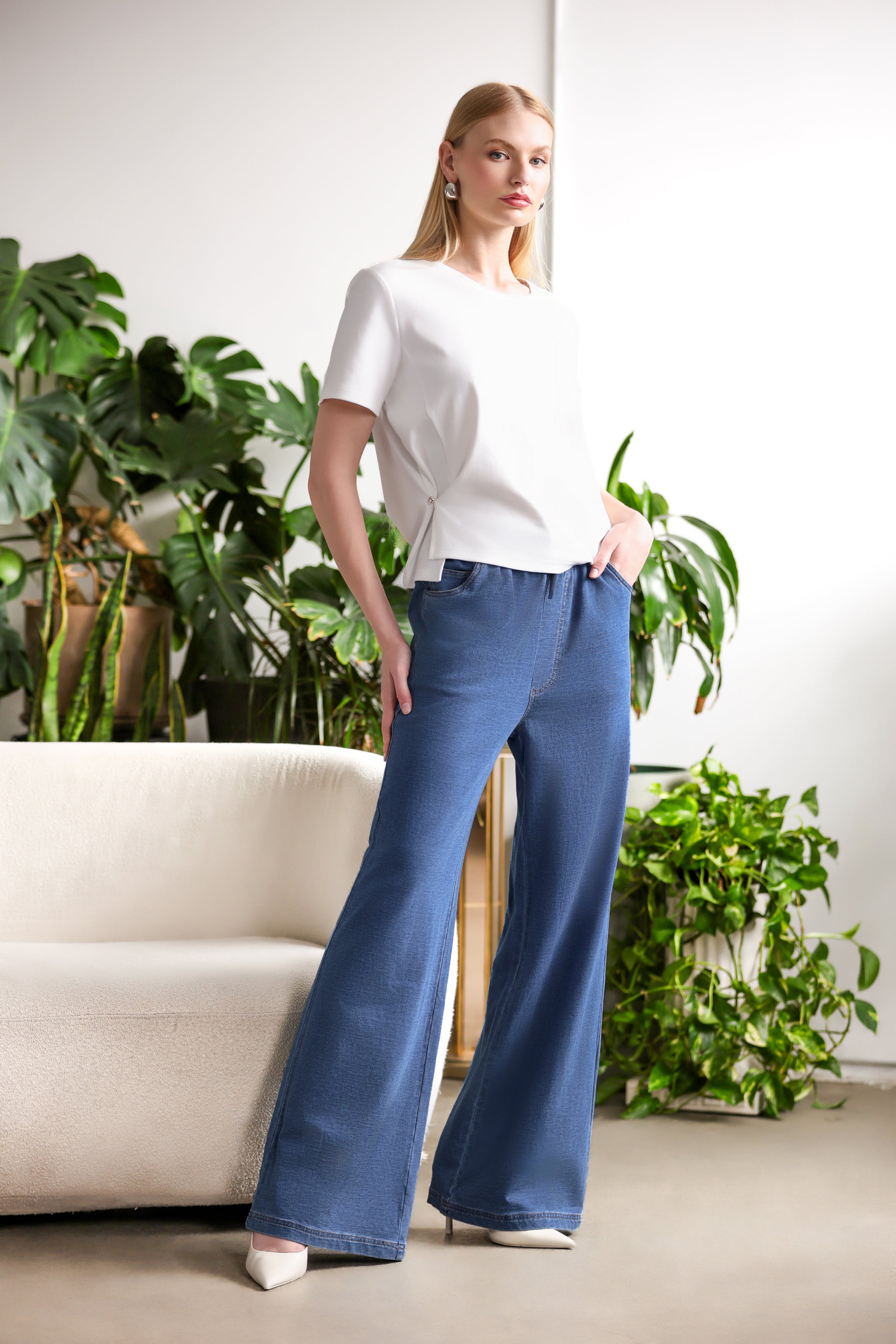 Model wearing a white short-sleeve asymmetric blouse with high-waisted wide-leg blue jeans and ivory pointed heels, styled indoors with green plants and a neutral sofa backdrop.