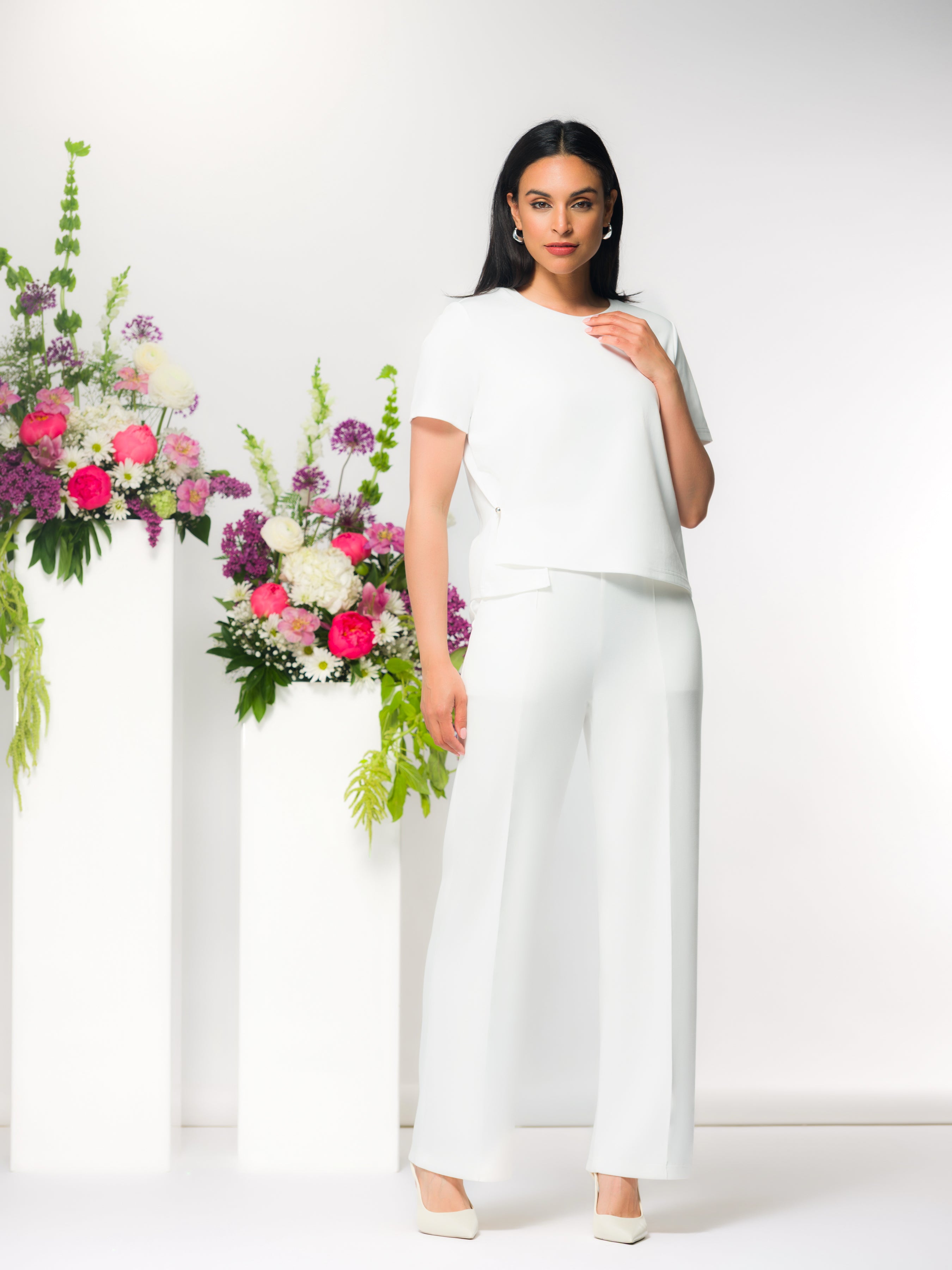 Model wearing a white short-sleeve asymmetric blouse with wide-leg white trousers and ivory heels, styled against a neutral studio background with floral arrangements.