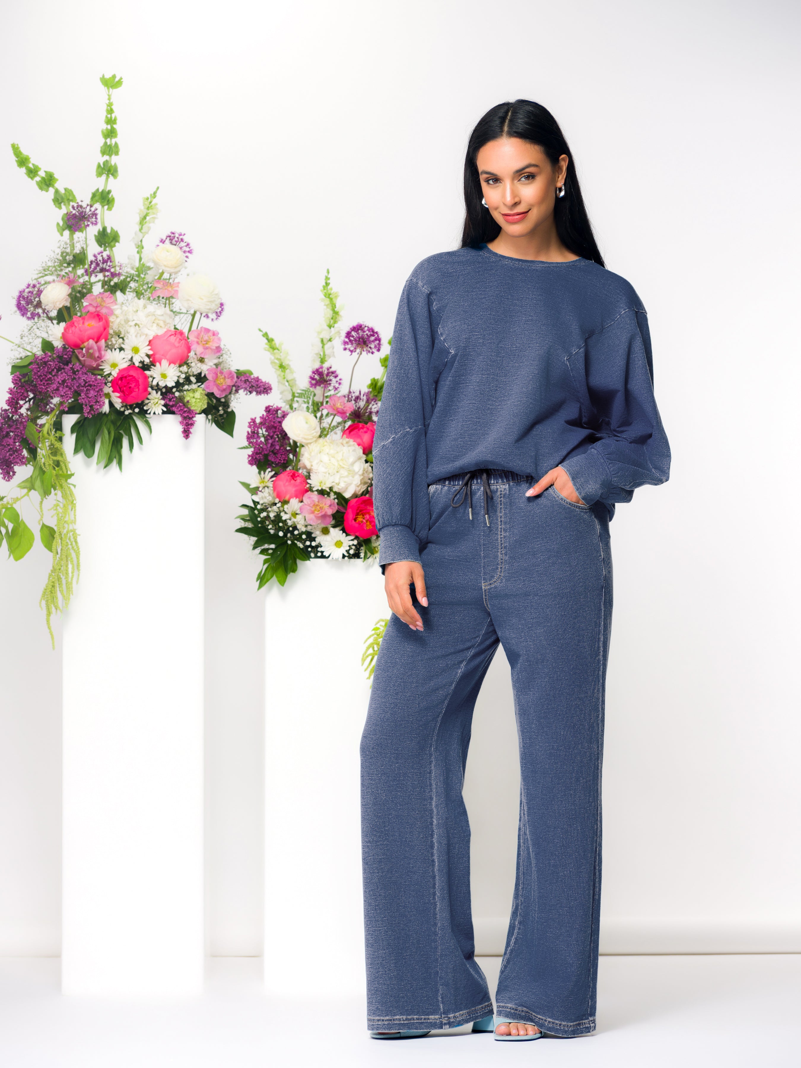Model wearing a blue relaxed-fit crewneck sweatshirt with dropped shoulders, styled with matching wide-leg pants in a bright studio setting.
