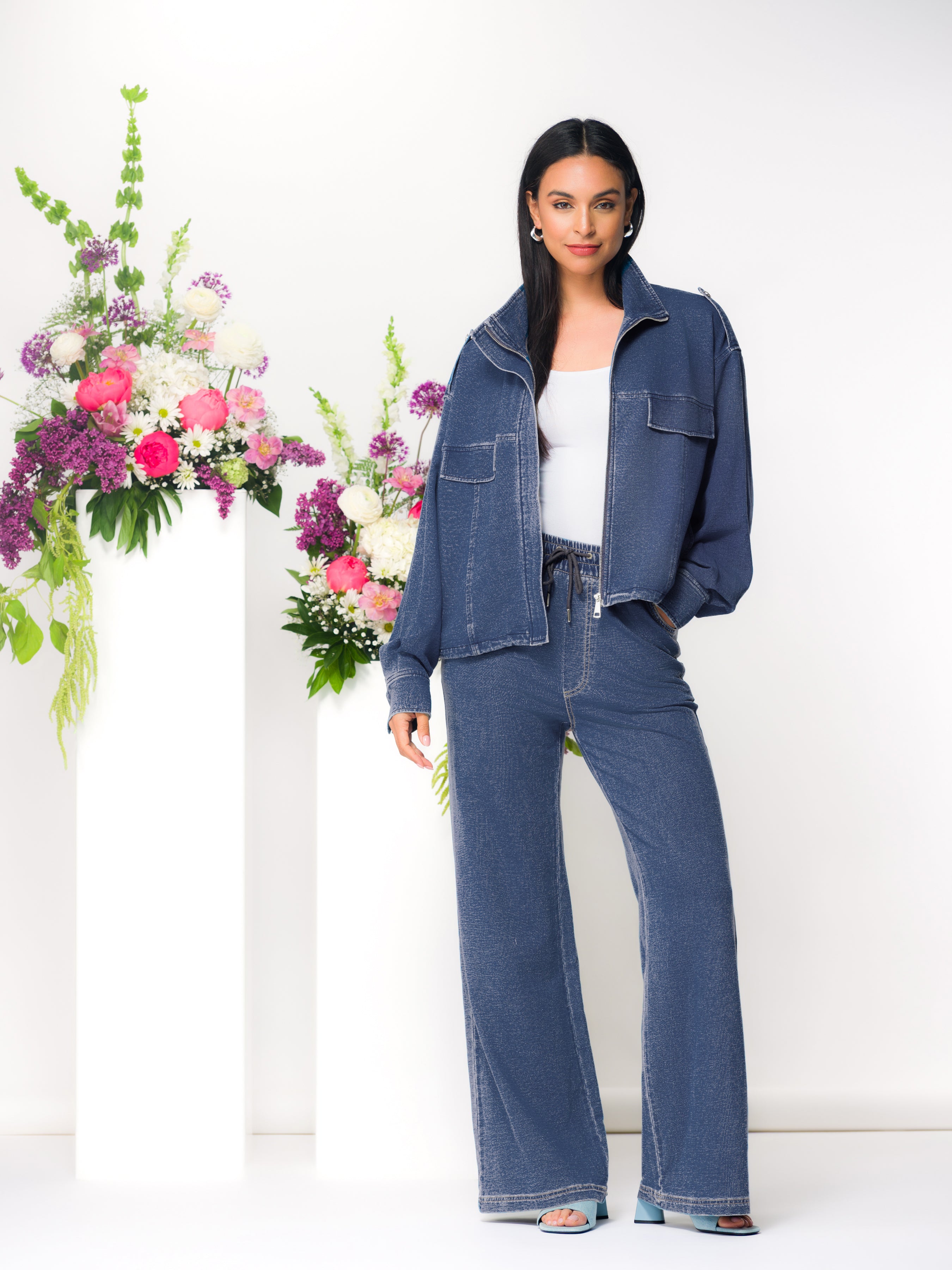 Model wearing a blue zip-up cardigan with matching wide-leg pants and a white top, standing beside a floral arrangement on white pedestals.