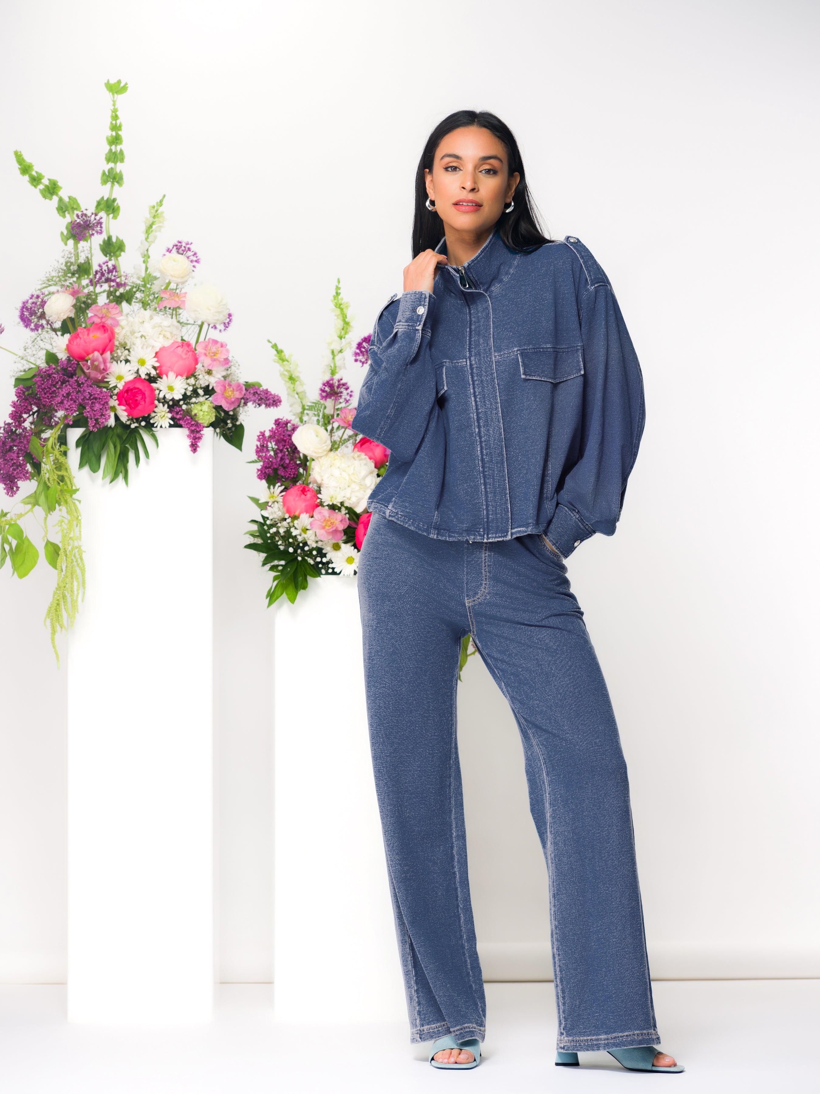 Model wearing a blue zip-up cardigan with matching wide-leg pants and a white top, posing with hand at collar next to floral display on white pedestals.