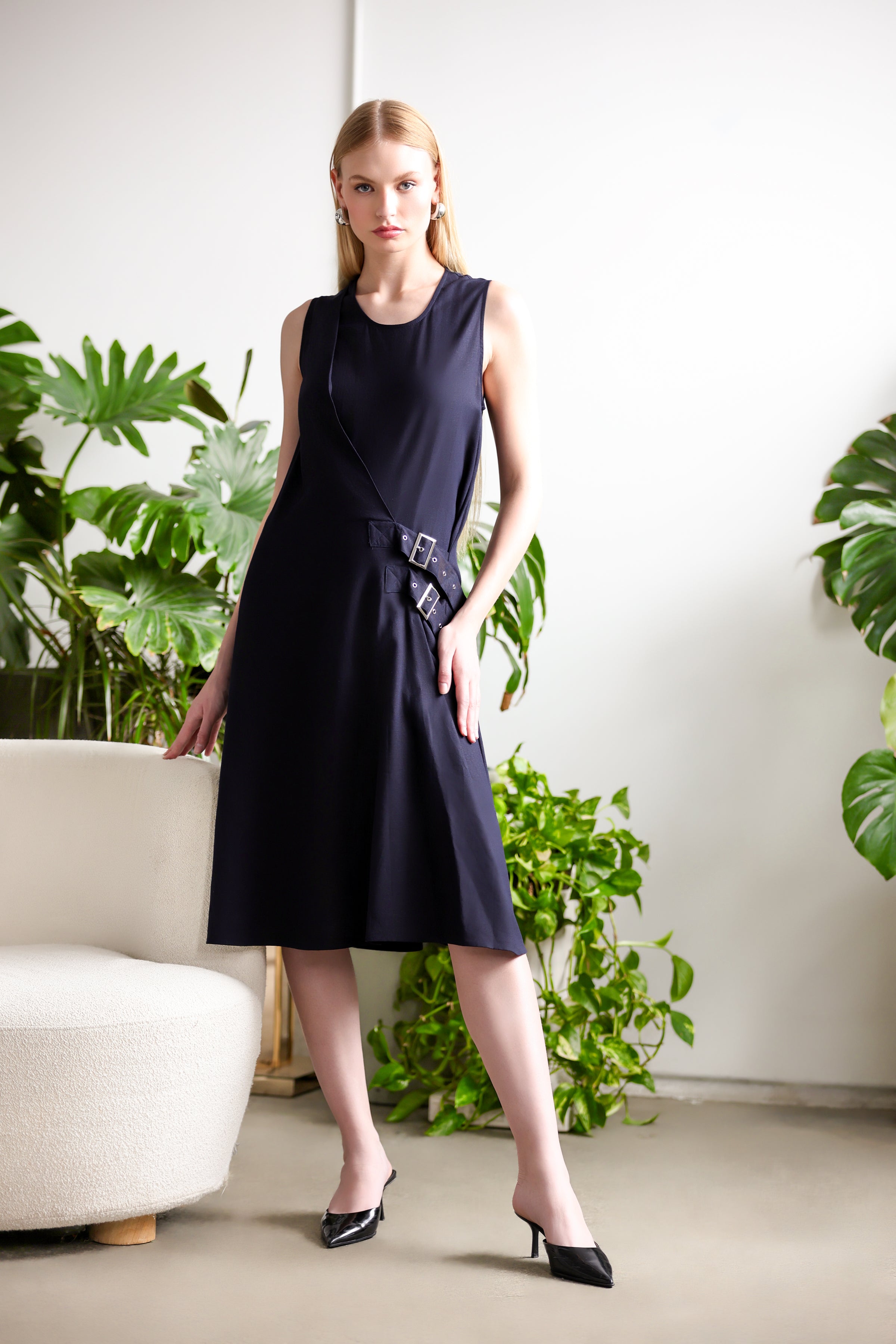 Model wearing a navy sleeveless midi A-line dress with side buckle detail, styled with black pointed heels, standing indoors beside a cream chair and green plants.
