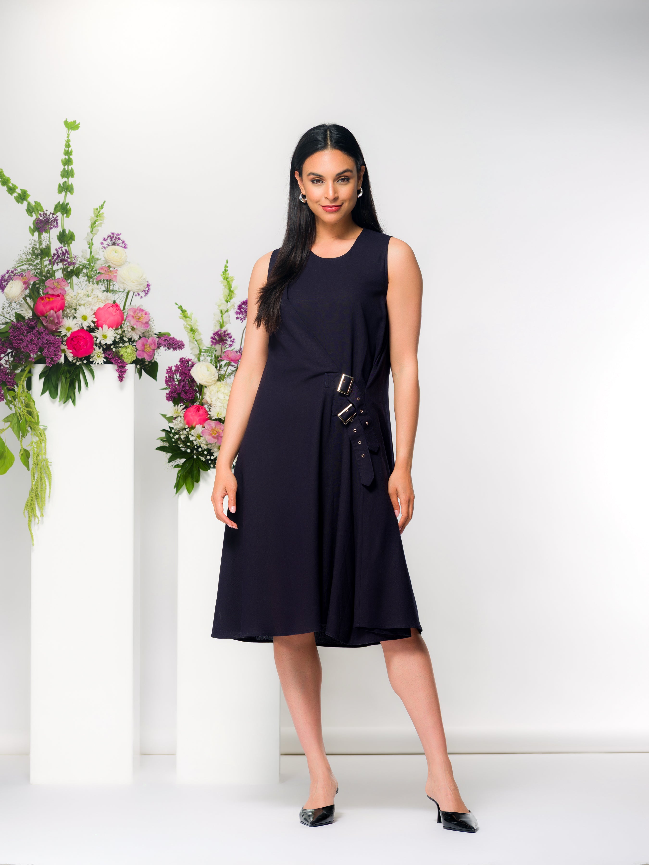 Model wearing a navy sleeveless midi dress with asymmetrical drape and side buckle accents, paired with black pointed heels, posed beside white pedestals with floral arrangements.