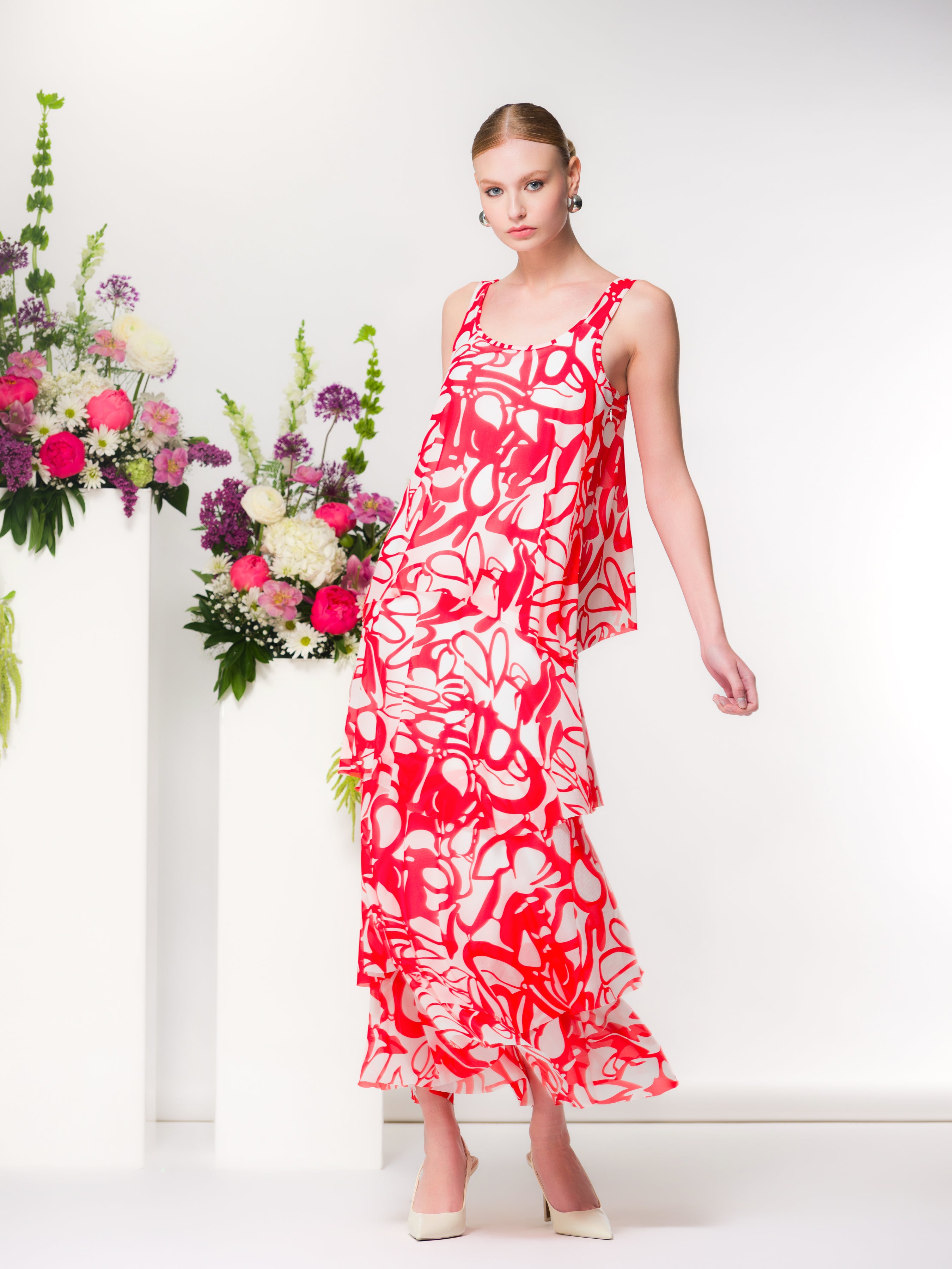 Sleeveless red and ivory abstract print tiered maxi dress with a flowy silhouette, styled with neutral pointed heels, shown on a model against a white studio backdrop with floral arrangements.
