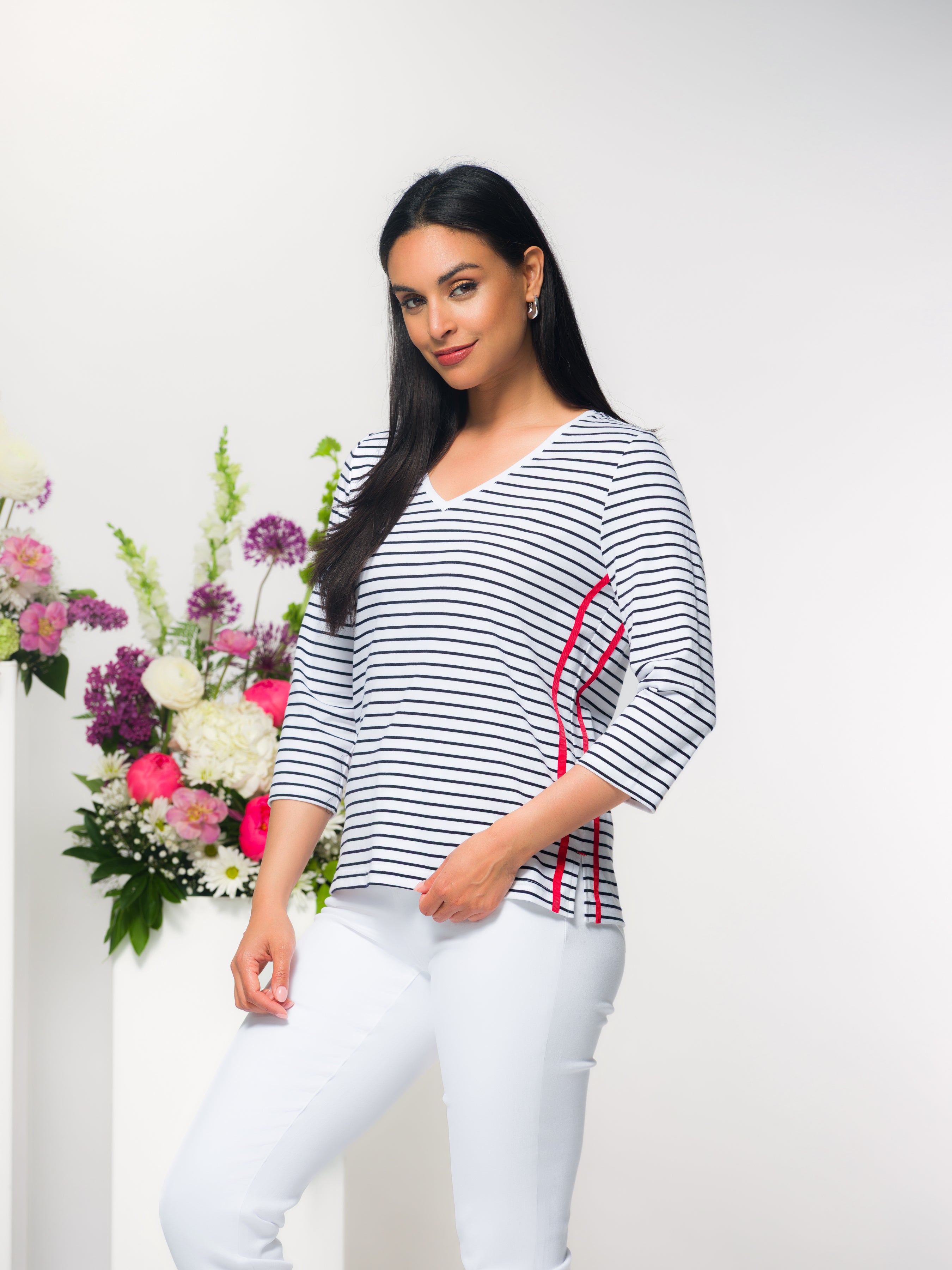 Woman modelling a black and white striped V-neck top with three-quarter sleeves and subtle side seam detailing, paired with light grey slim-fit pants, standing in a studio setting with a floral arrangement in the background.