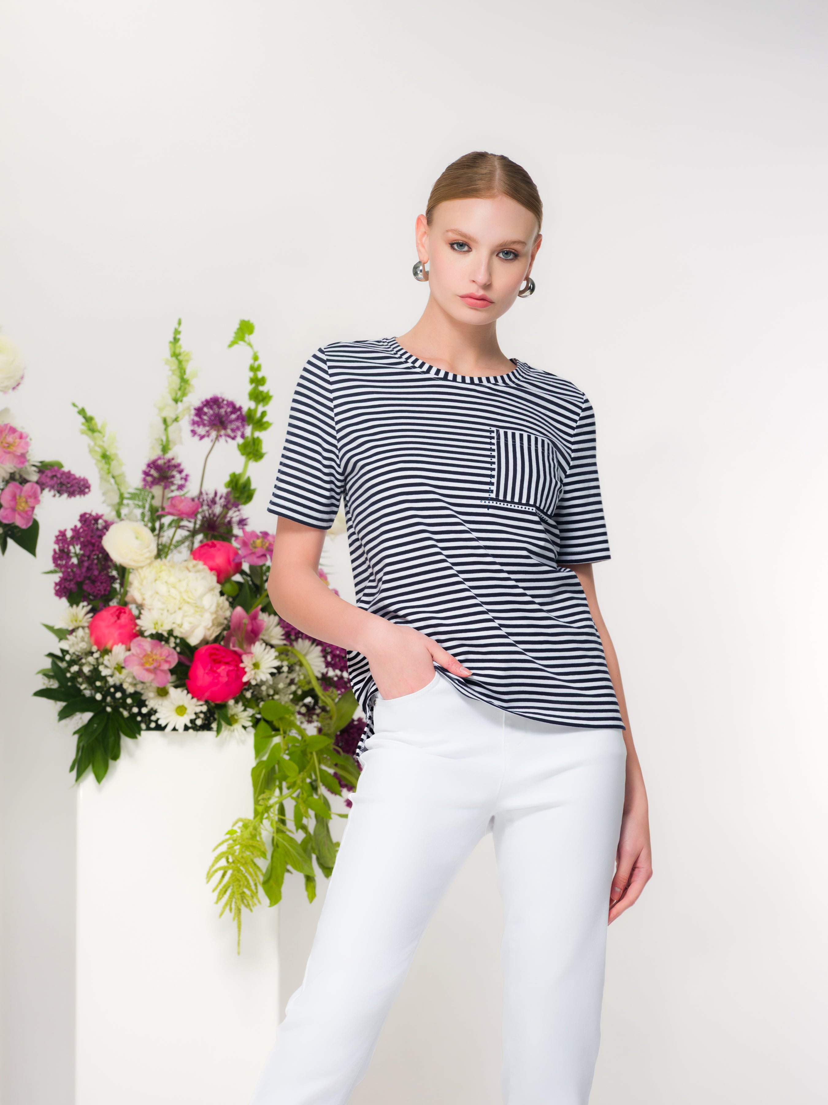 Model wearing a navy and white striped short sleeve cotton tee with chest pocket, paired with light grey trousers, studio background with floral arrangement.