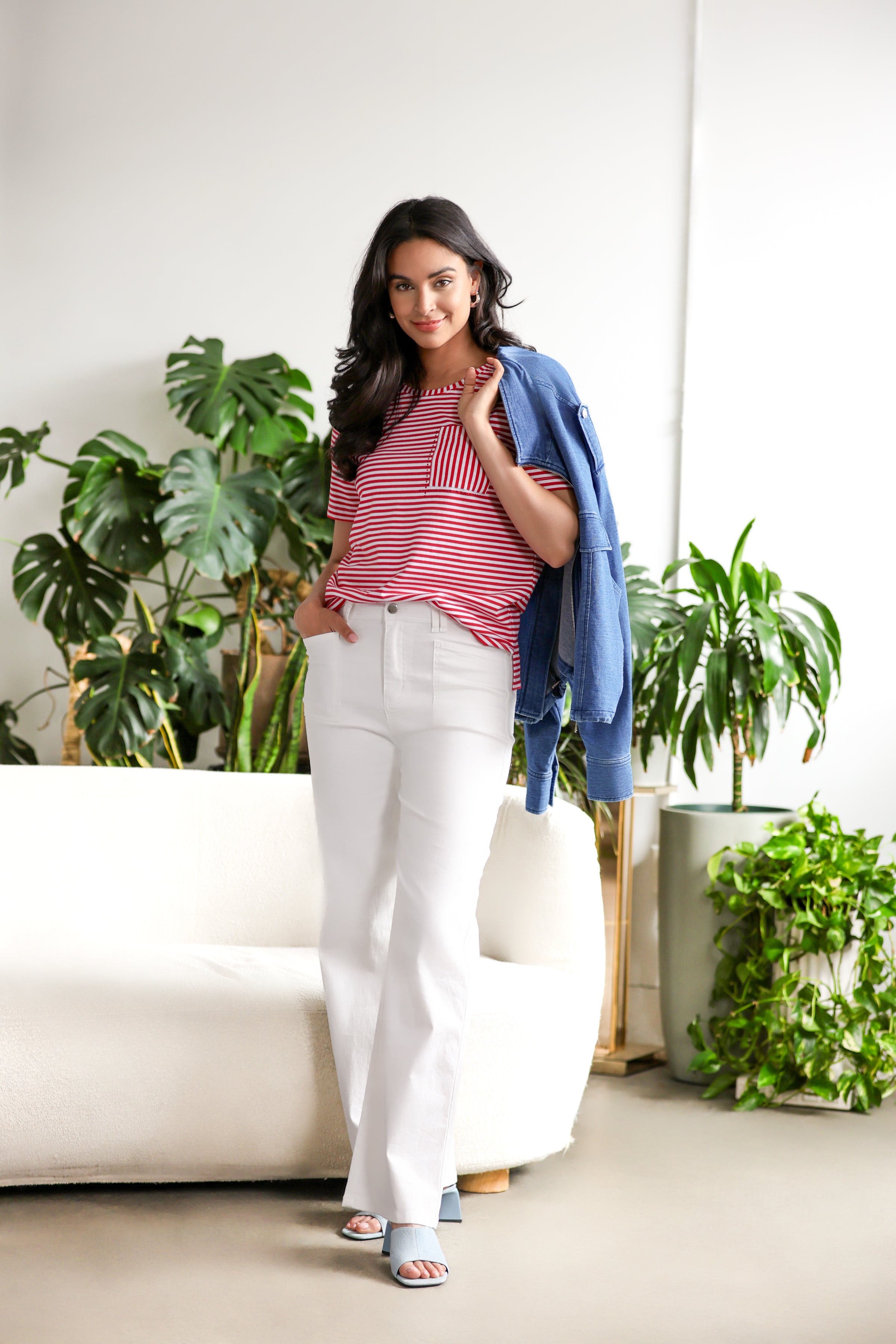 Model wearing a red and white striped short sleeve cotton tee with chest pocket, paired with white wide-leg pants and denim jacket, styled in a bright indoor setting with green plants.